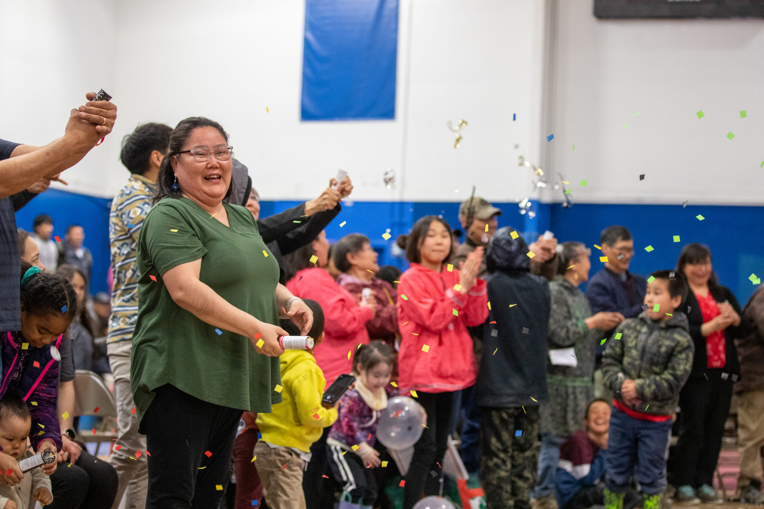 A crowd of people pop confetti canisters in a school gymnasium.