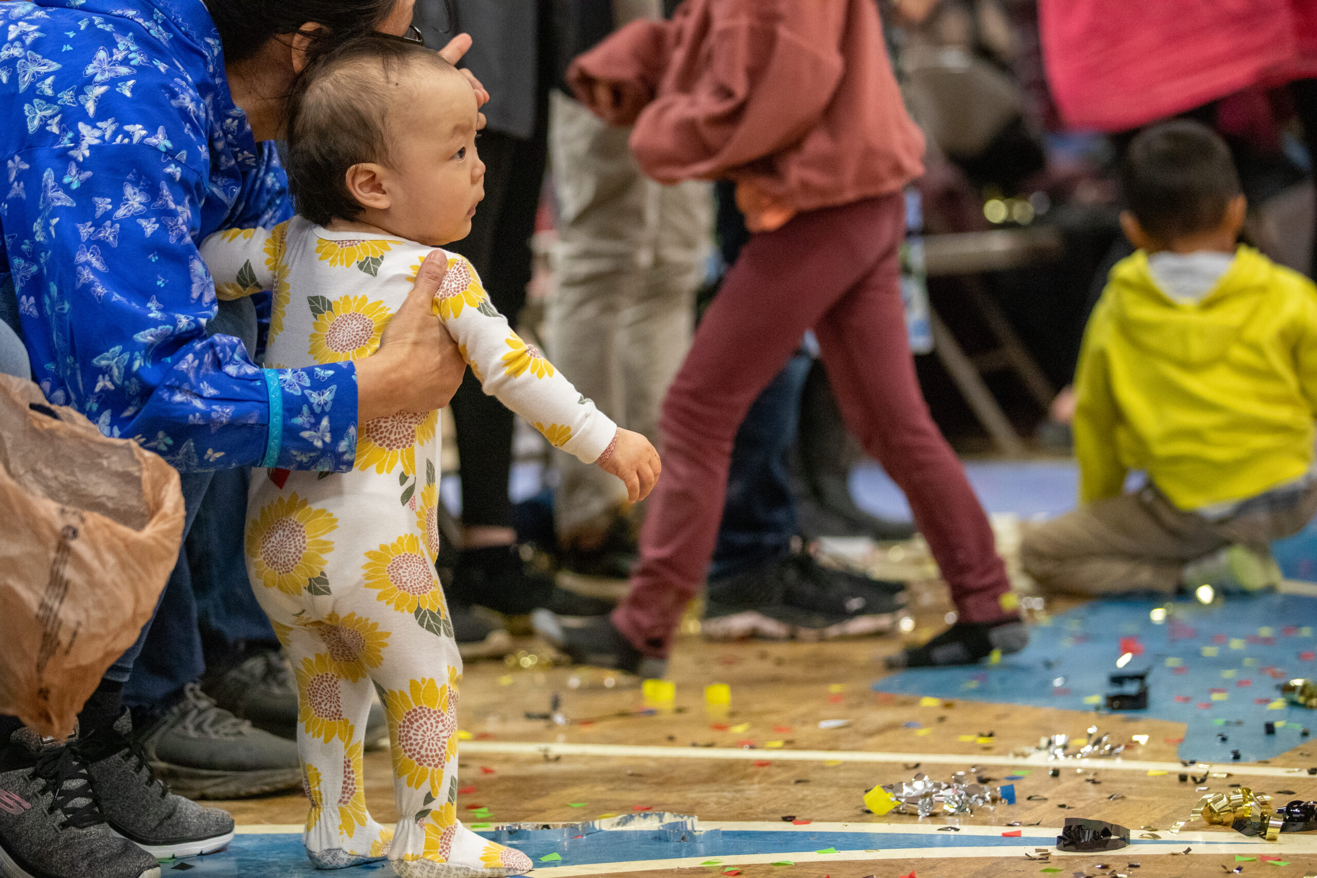 A baby stands on a gym floor surrounded by confetti.