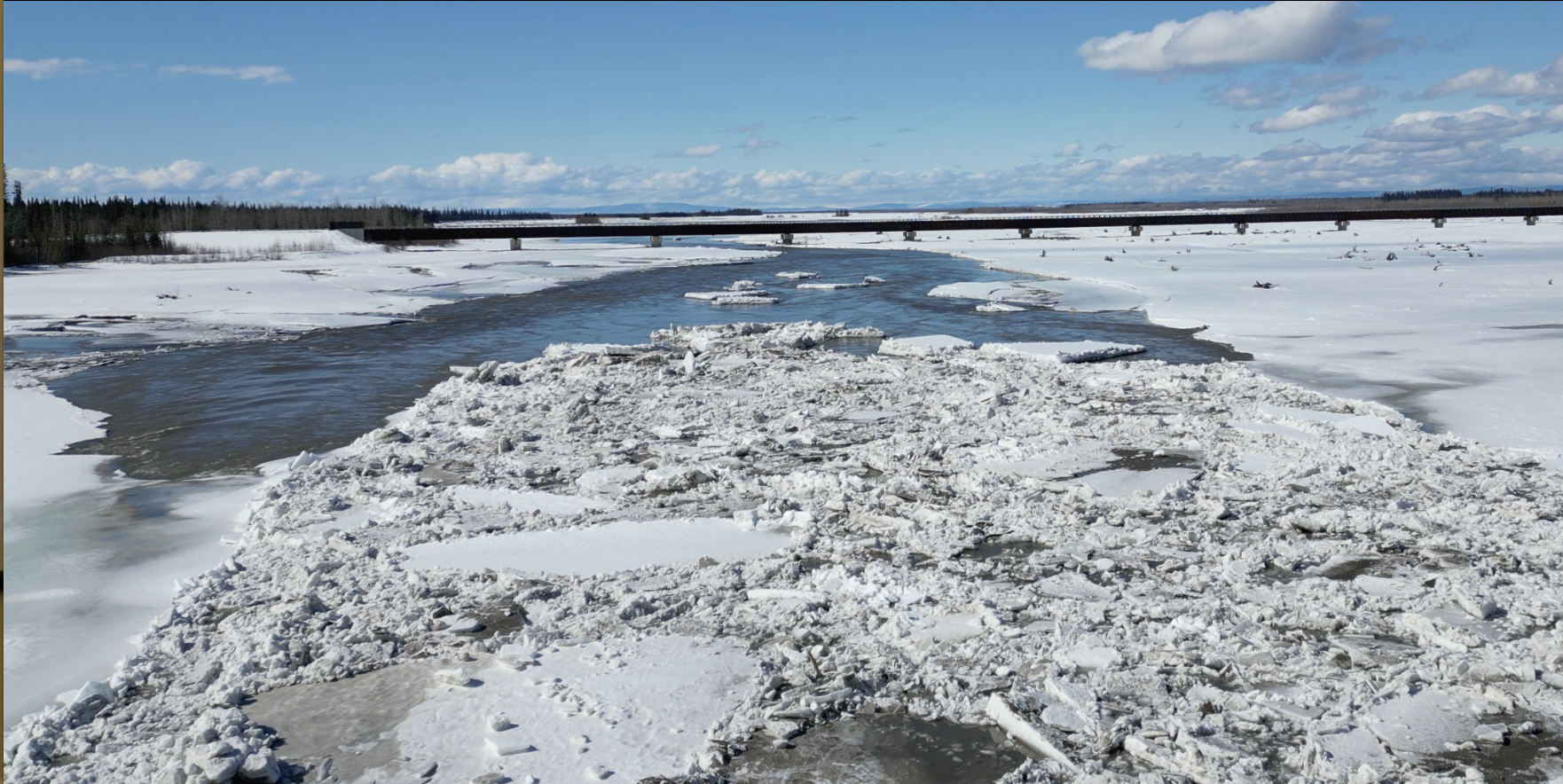 A river mostly covered in ice on a sunny day with a bridge in the distance.