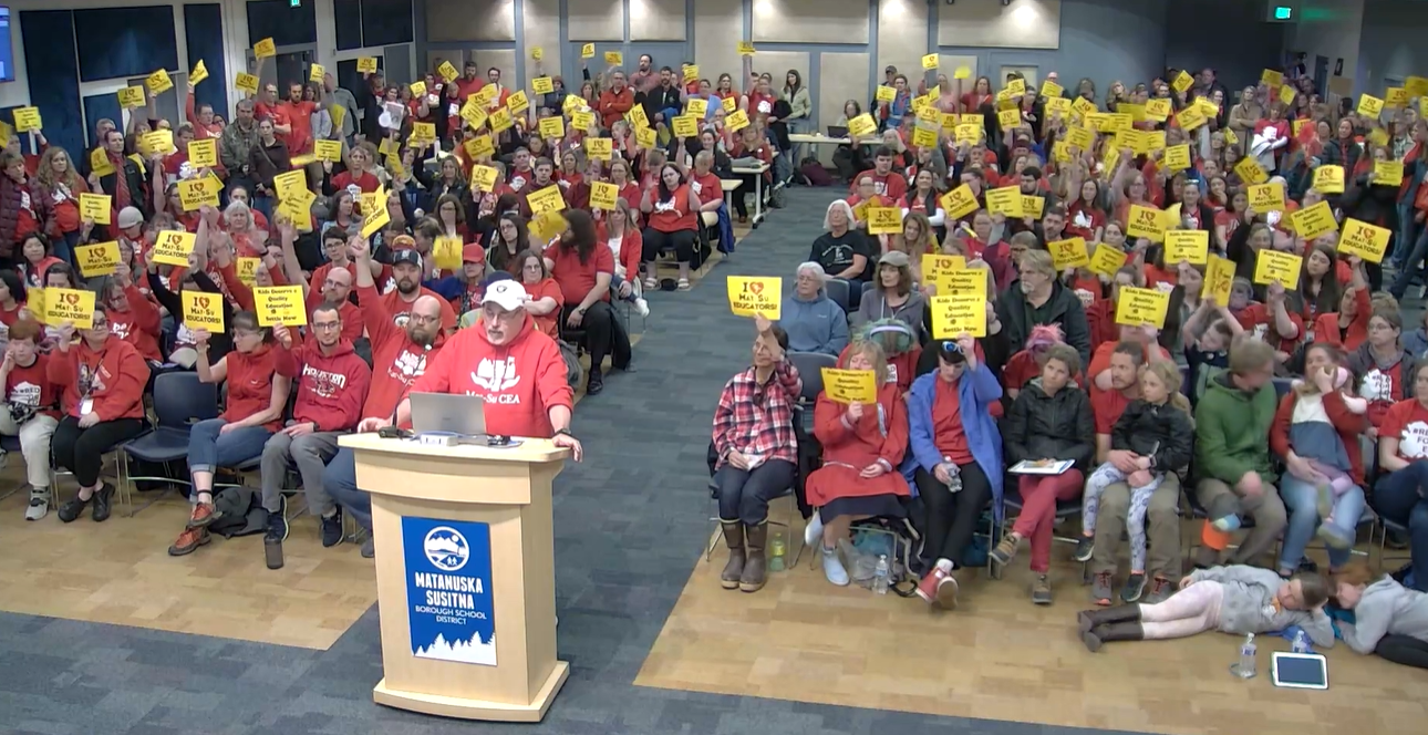 A man in red speaks in a crowd of people wearing red, waiving yellow signs.