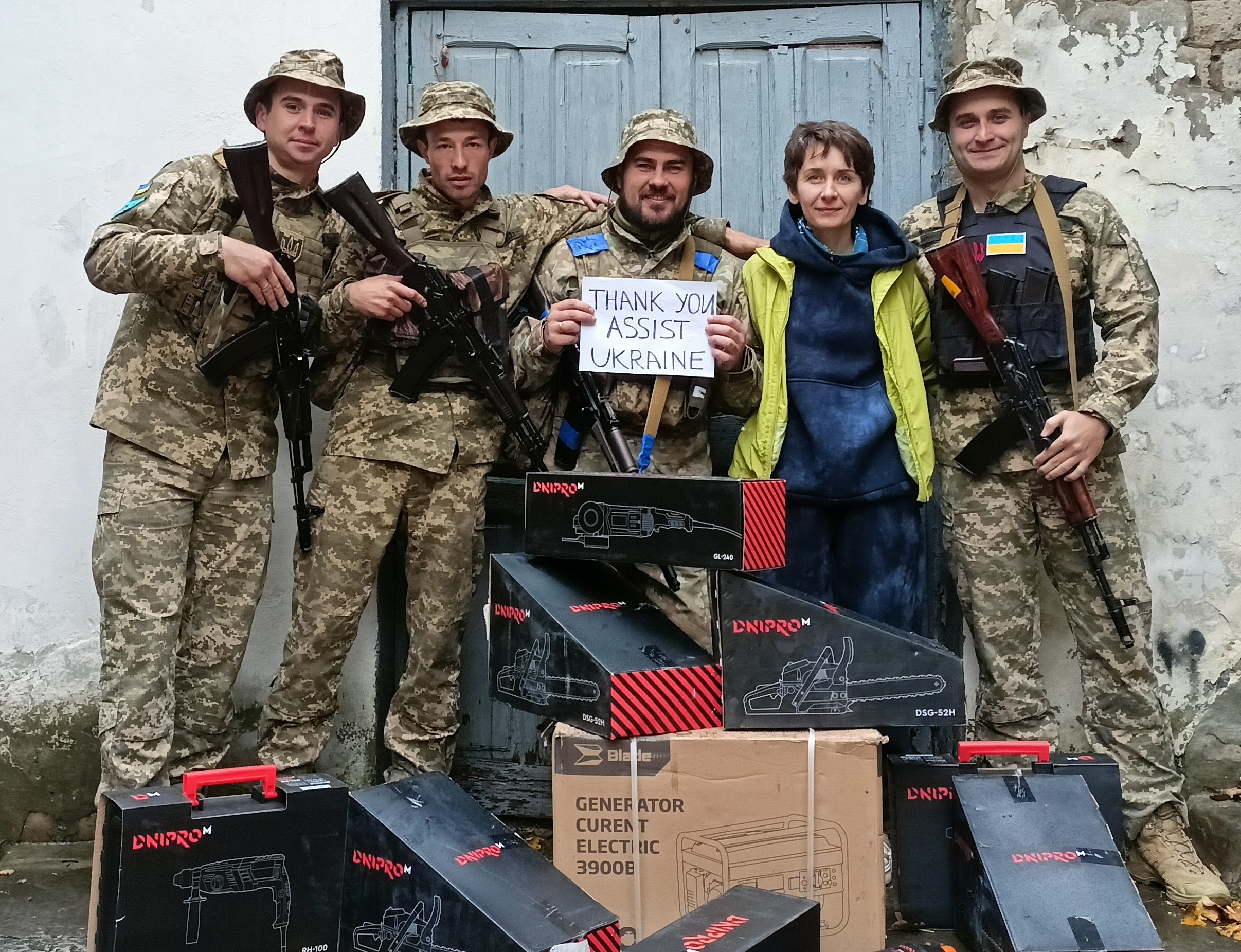 Four men in camouflage uniforms, each holding a rifle, pose for a photo with a woman in civilian clothes behind a pile of boxes. One is holding a sign that says, "Thank you, Assist Ukraine." 