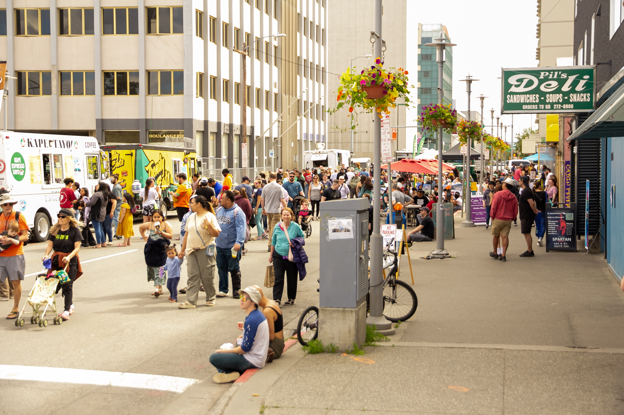 Crowds of people walk past food trucks and vendors along a city street on a summer afternoon.