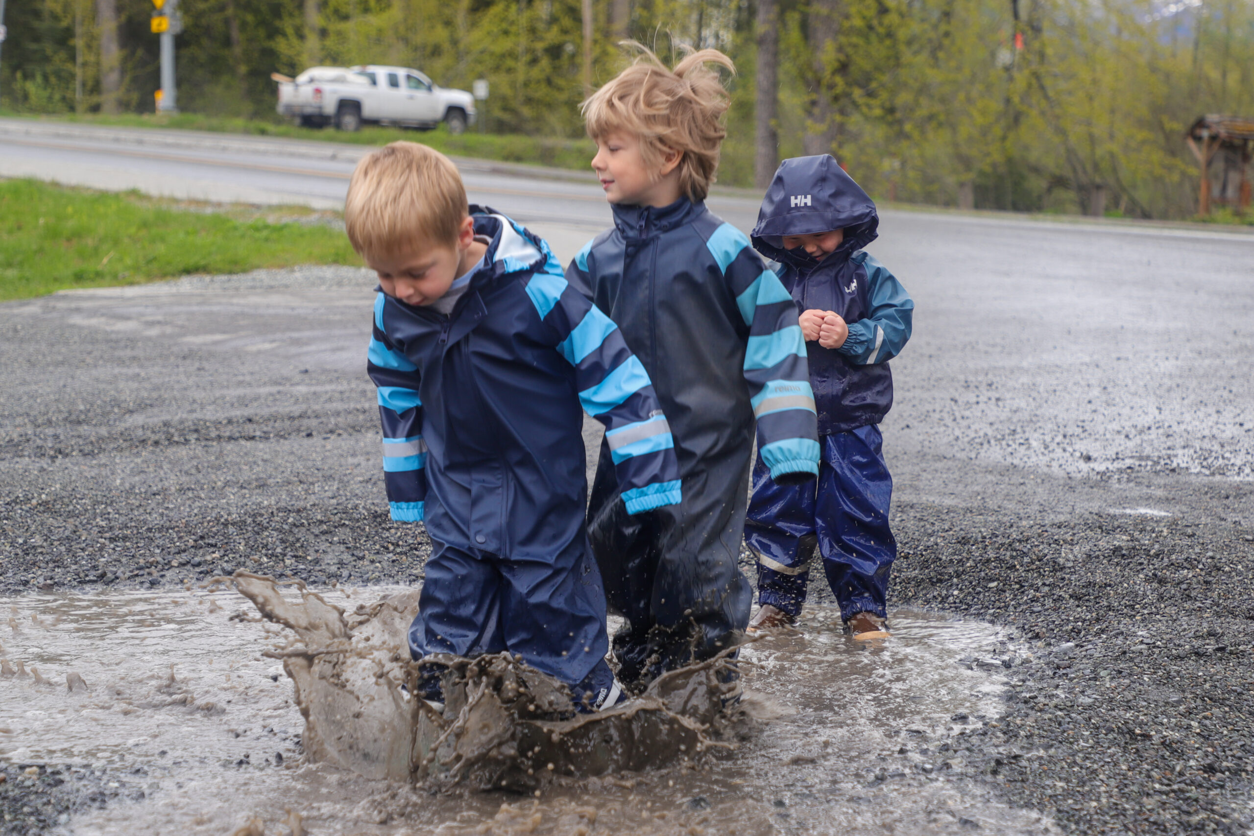 Three children in blue rain gear jump in a puddle in a gravel parking lot.
