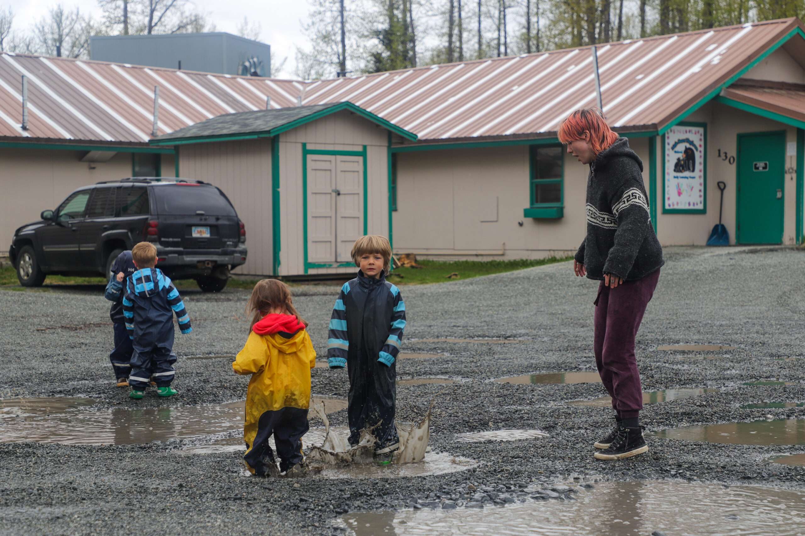 A person with dyed pink hair stands in a parking lot with four children who are splashing around in puddles.