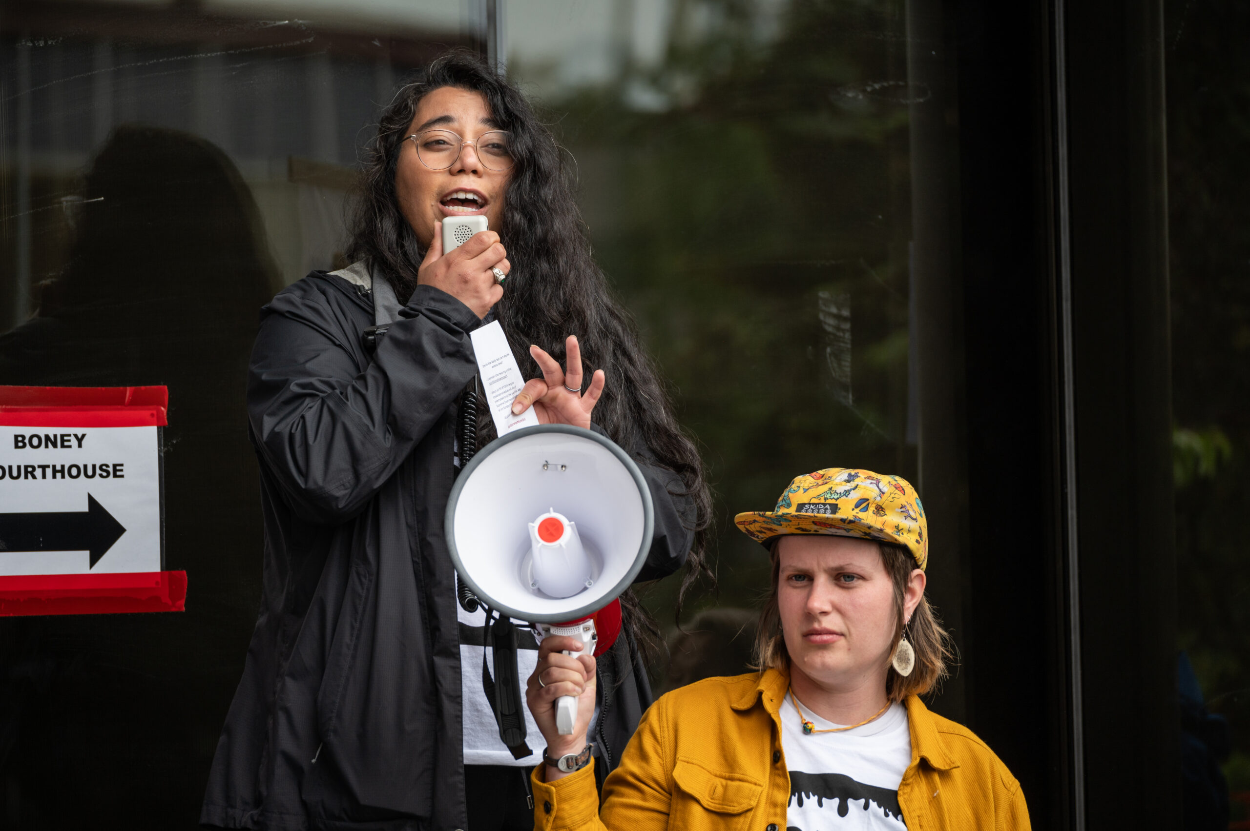 A woman with a black jacket on speaks into a megaphone, held by a person in a yellow shirt.