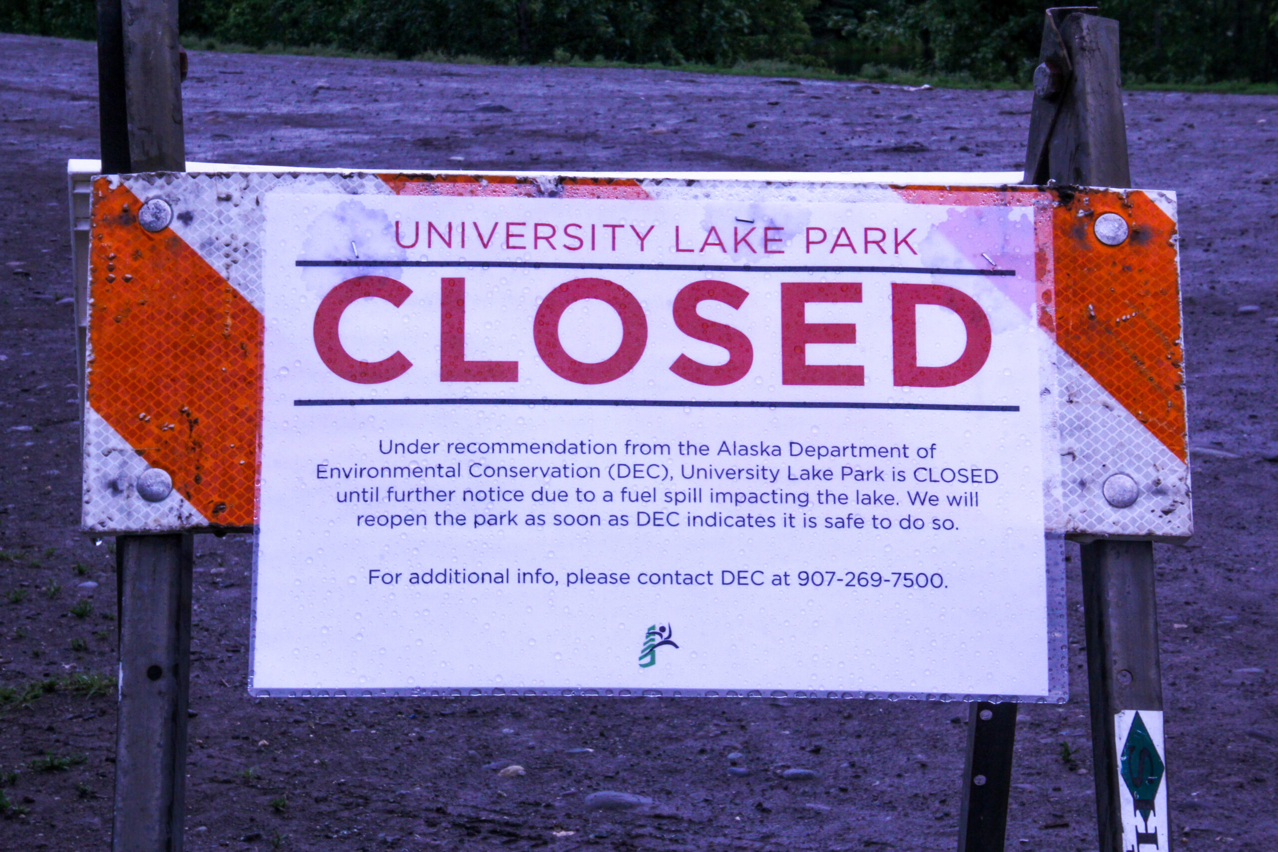 A sign that reads "University Lake Park CLOSED. Under recommendation from the Alaska Department of Environmental Conservation (DEC), University Lake Park is CLOSED until further notice due to a fuel spill impacting the lake. We will reopen the park as soon as DEC indicates it is safe to do so. For additional info, please contact DEC at 907-269-7500."