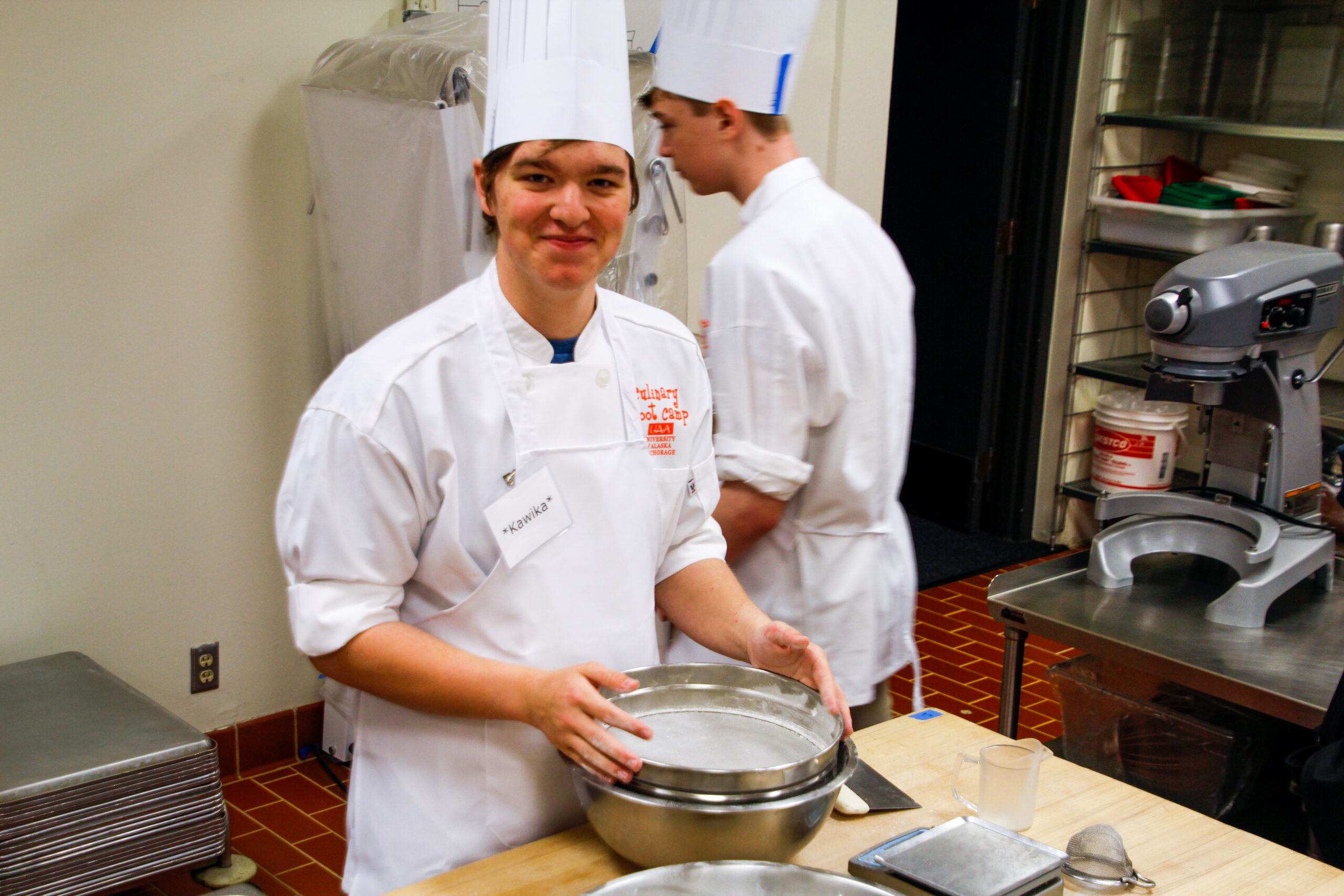 A teenager in a white chef's coat and hat stands at a kitchen counter, with a mixing bowl and sifter.