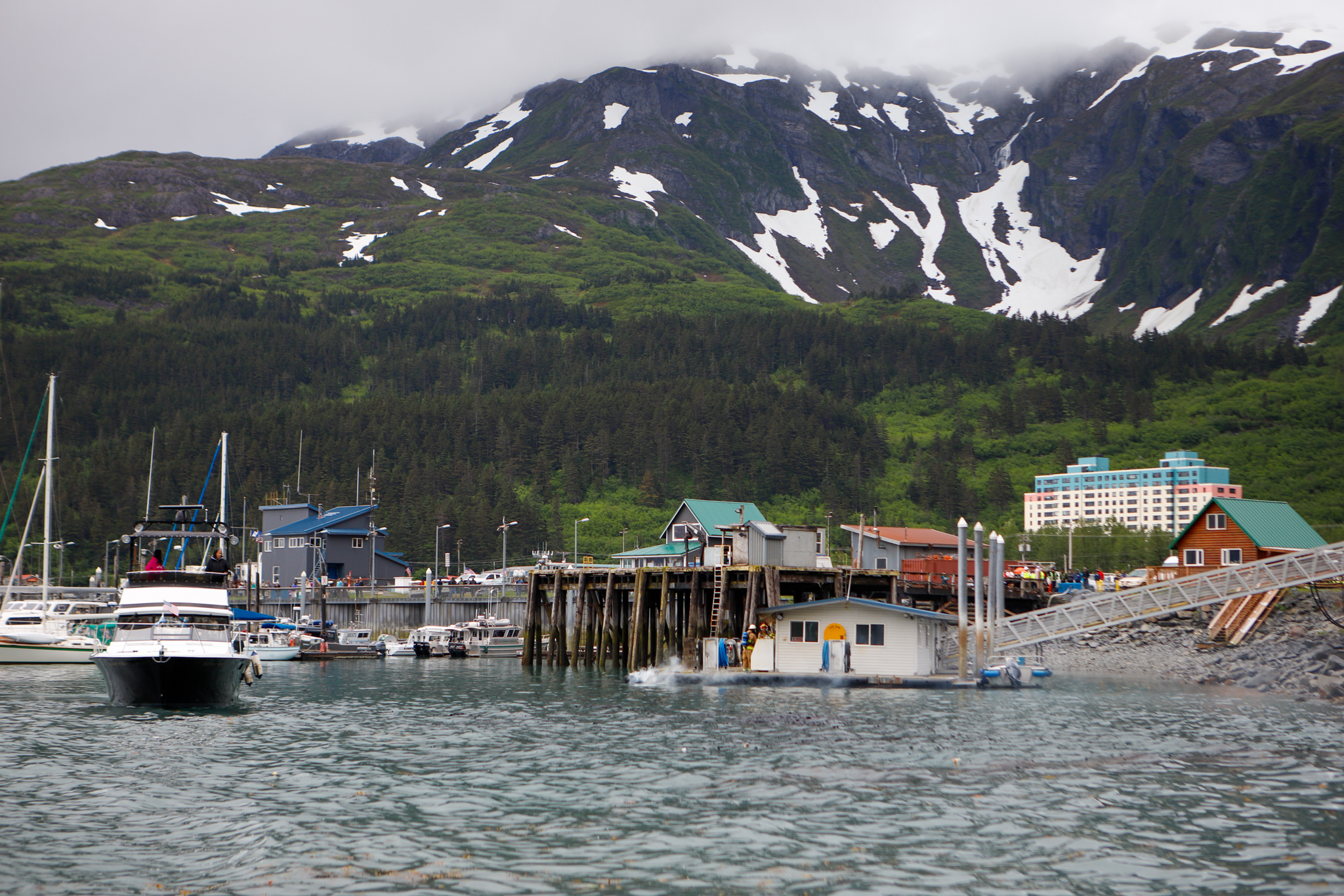 A wideshot of a boat harbor in front of a snow-capped mountain with a small dock fire smoking.