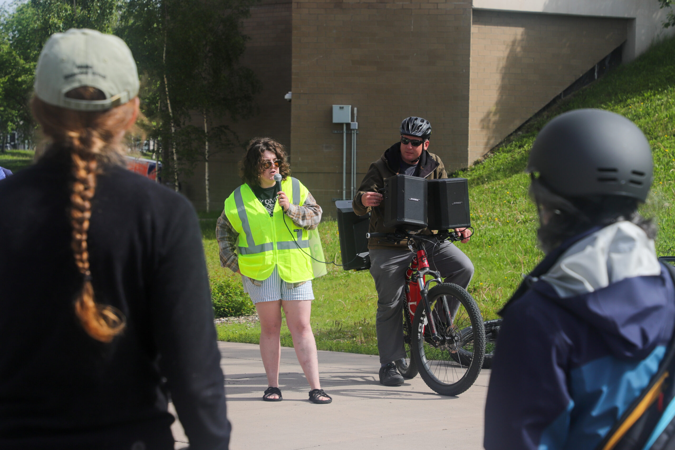 A woman in a high vis vest with a microphone standing next to a man on a bike with speakers attached to it.