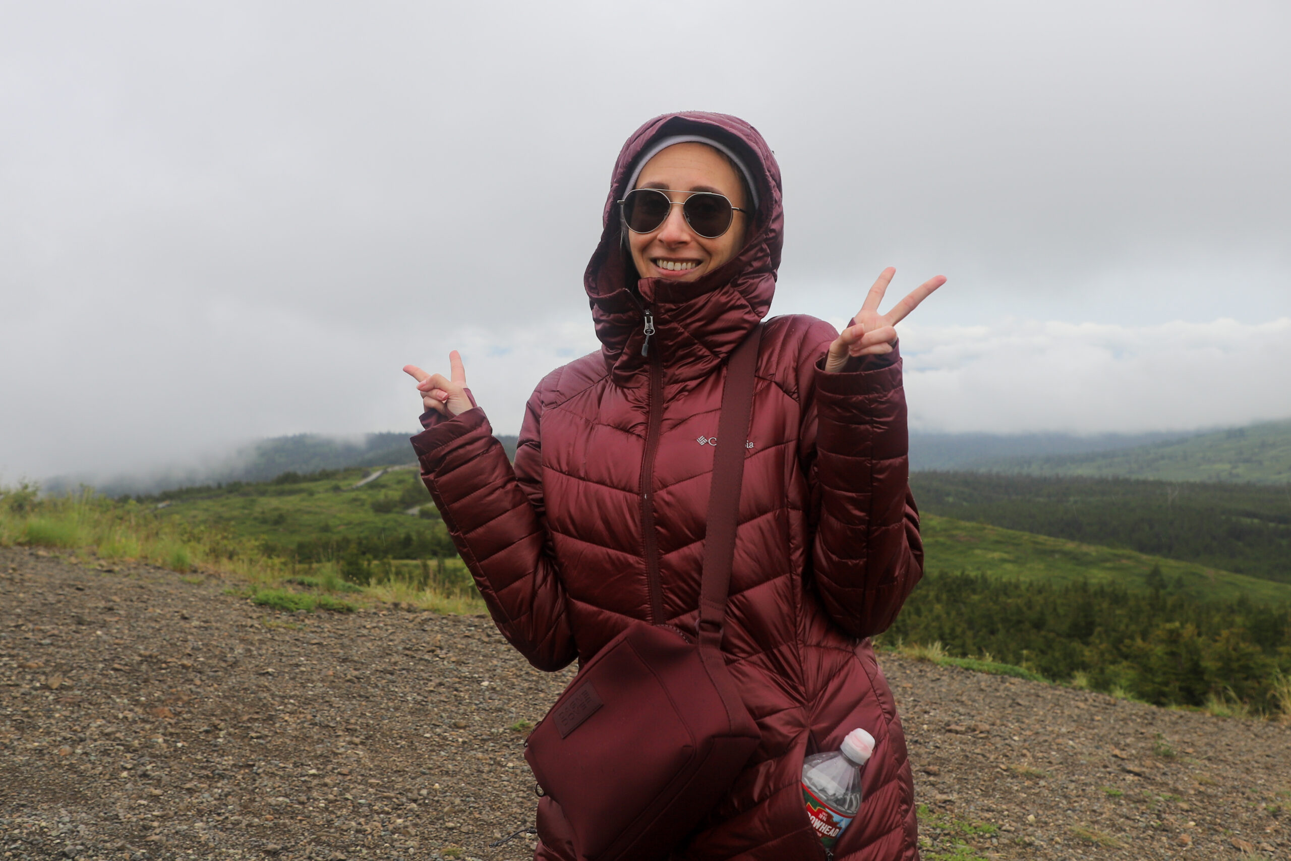 a woman in a maroon hooded jacket and sunglasses makes double peace signs on a mountain trail.