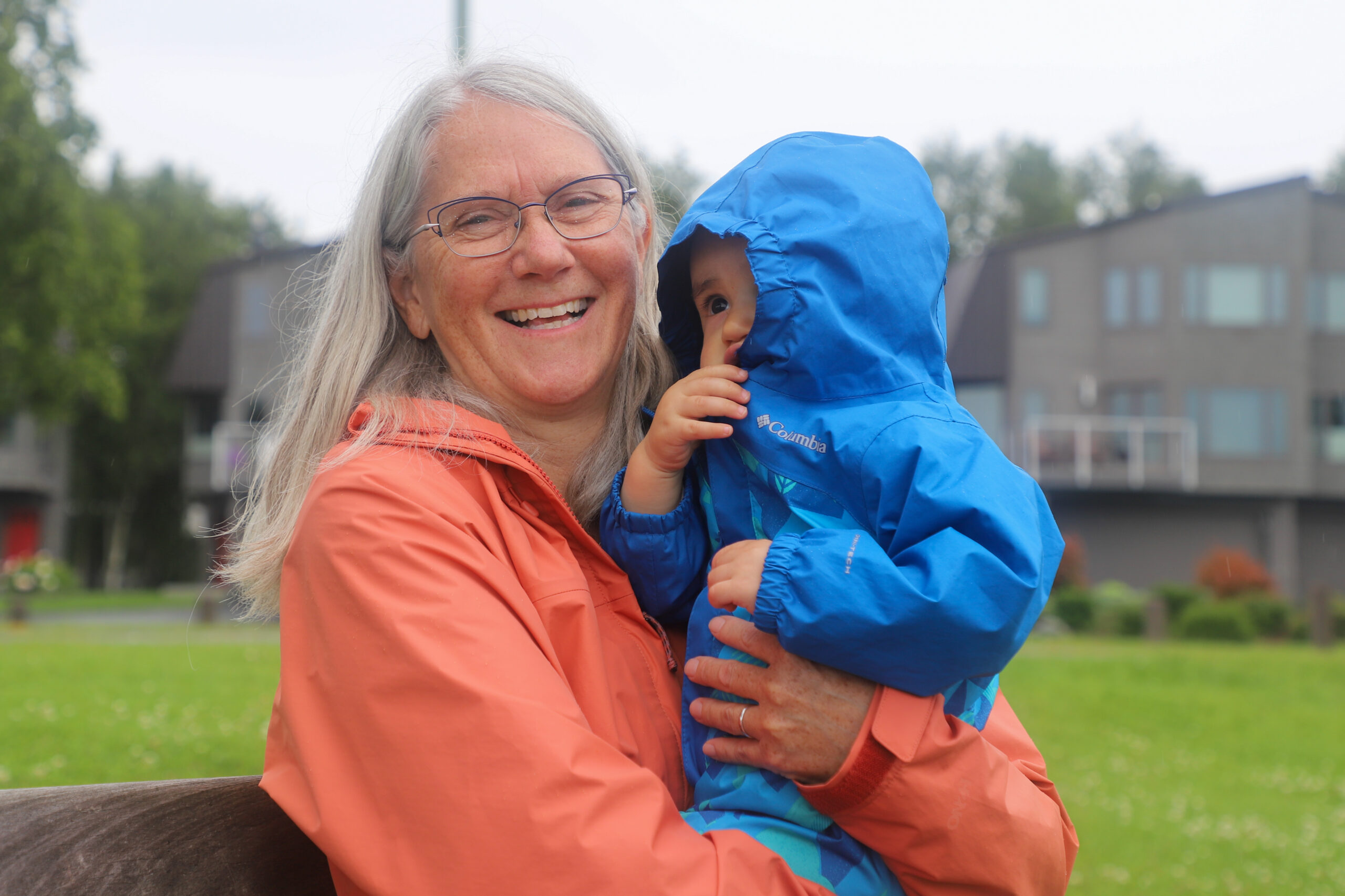 A woman with grey hair in a pink raincoat holds a child wearing a blue raincoat