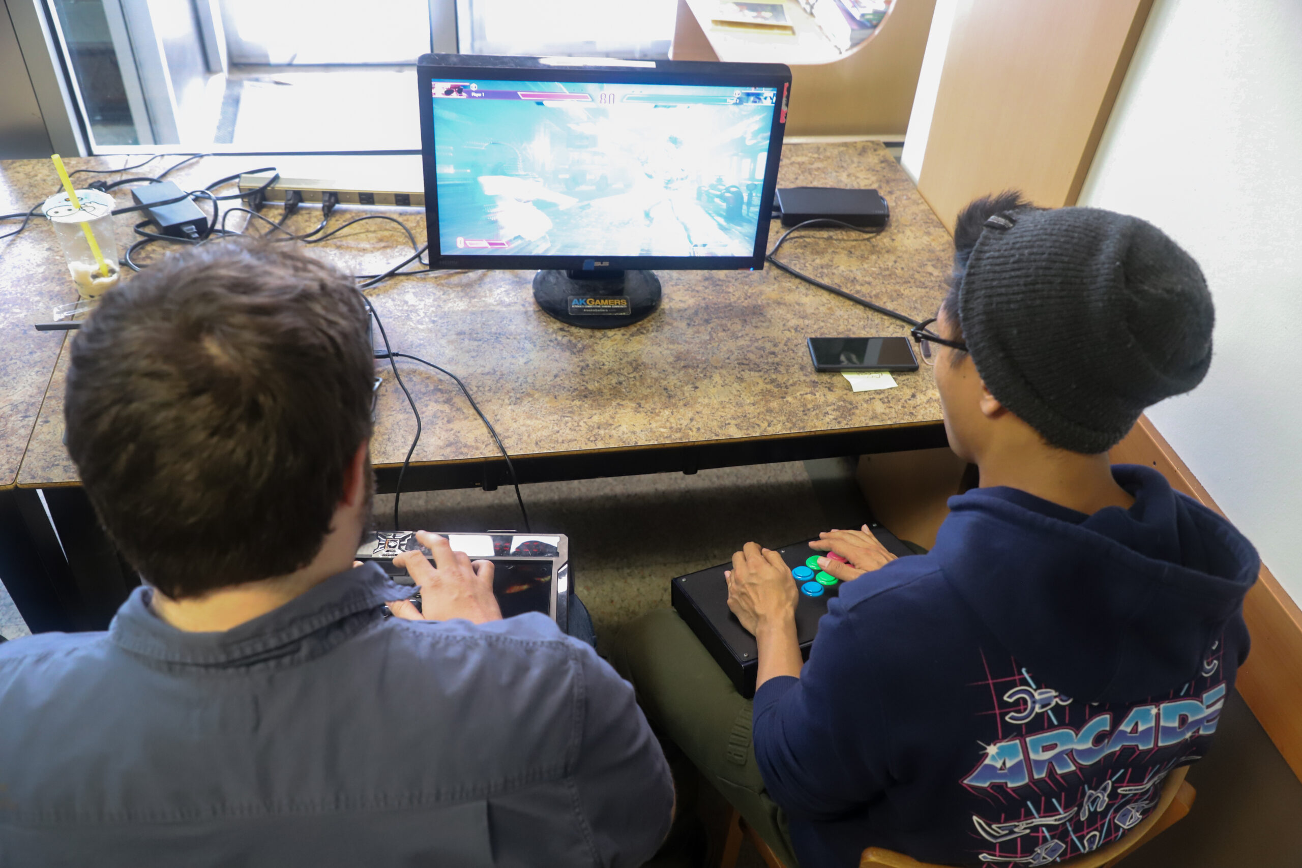 Two people sit at a table and play a video game. They have boxy controllers with buttons and levers on their laps.