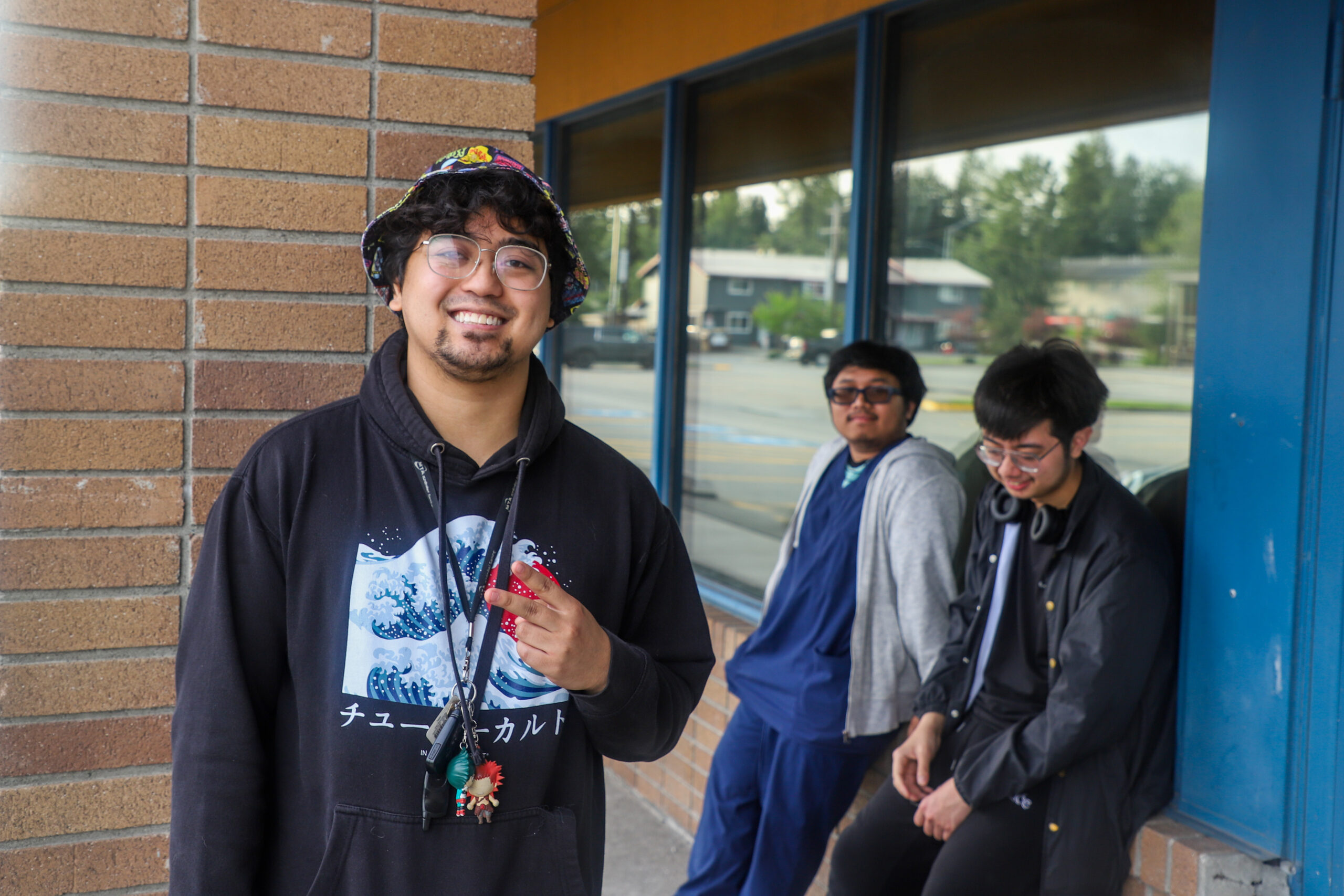 A man stands outside of a strip mall, wearing a colorful bucket hat and a lanyard with keychains on it. He throws a peace sign. Two people lean on the wall behind him.