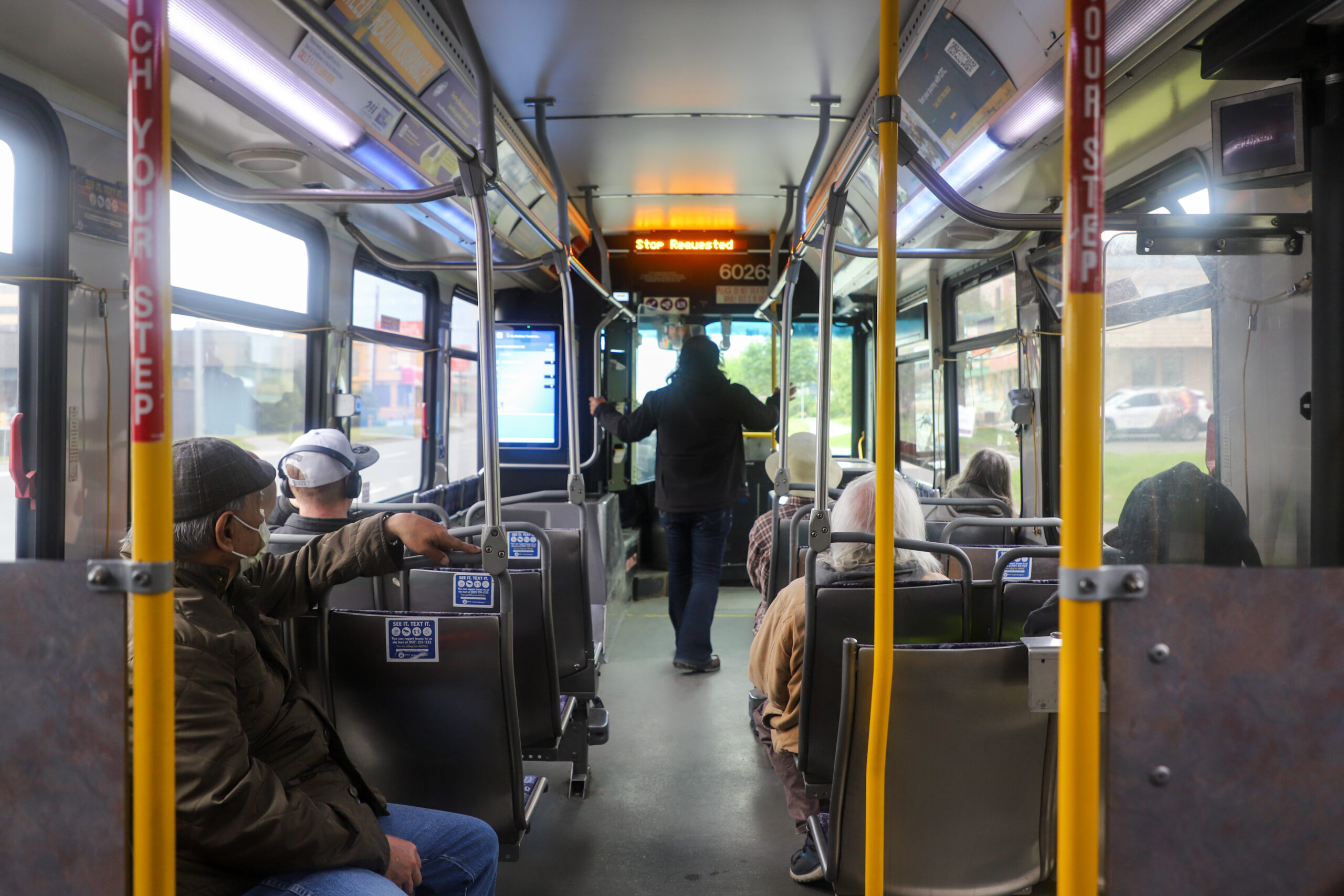 The inside of a bus. One passenger stands in the middle, walking to the front door. People sit on either side.