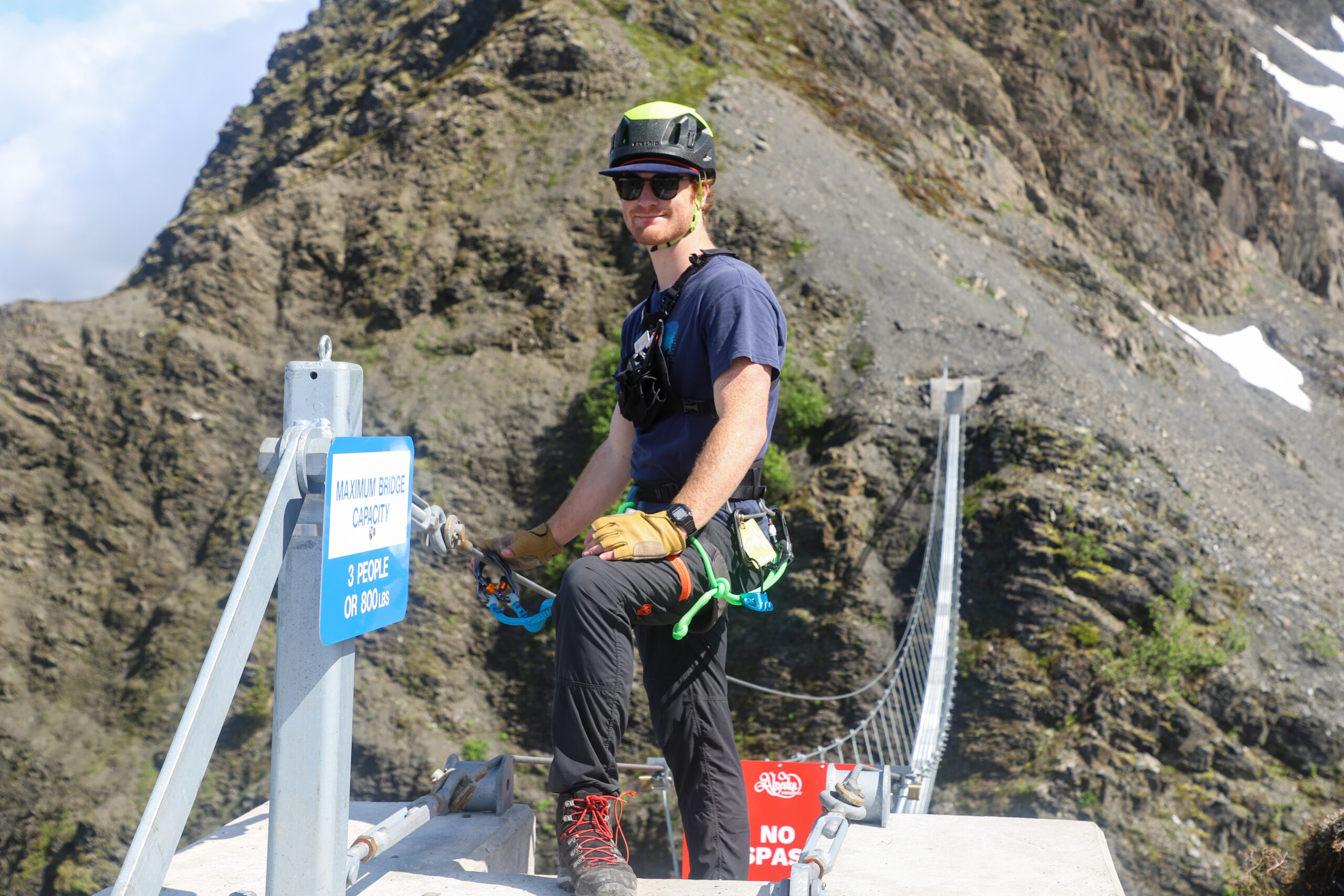 a person with a helmet and climbing harness waiting at the end of a long suspension bridge across a mountainside.