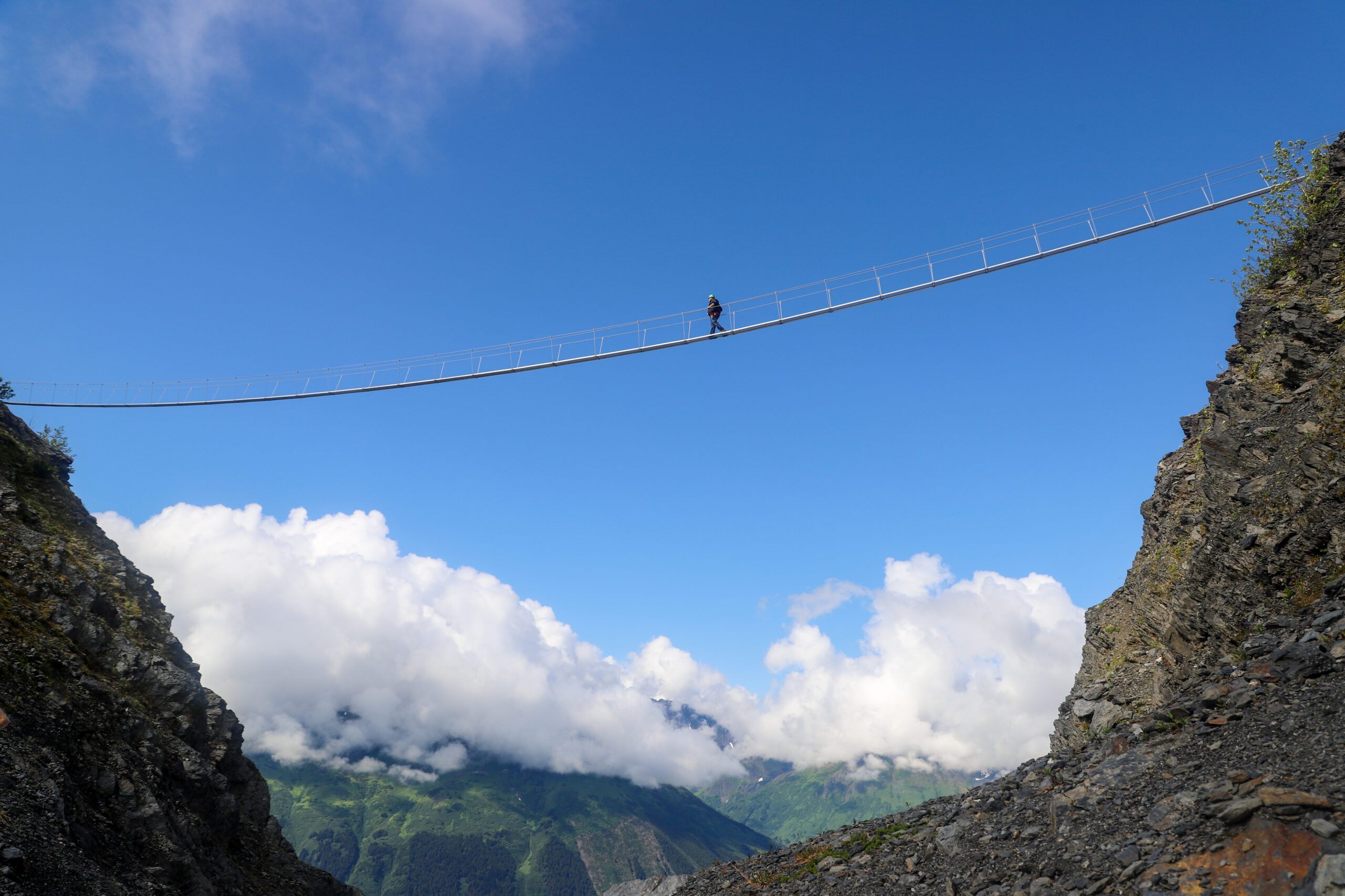 A person walks across a narrow bridge high above the ground, suspended between two cliff sides.