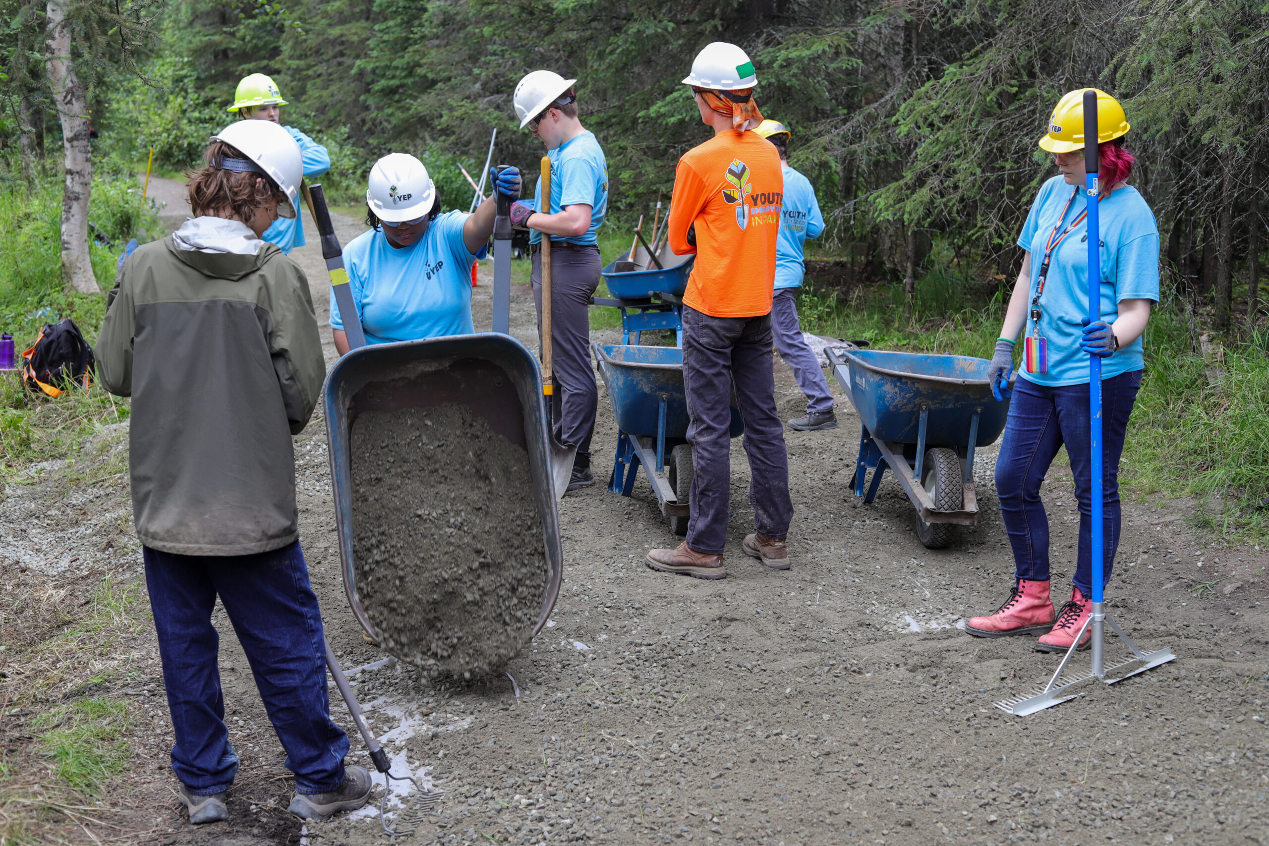 A person in a hard hat dumps a wheelbarrow full of gravel onto the ground. Others stand next to them with rakes.