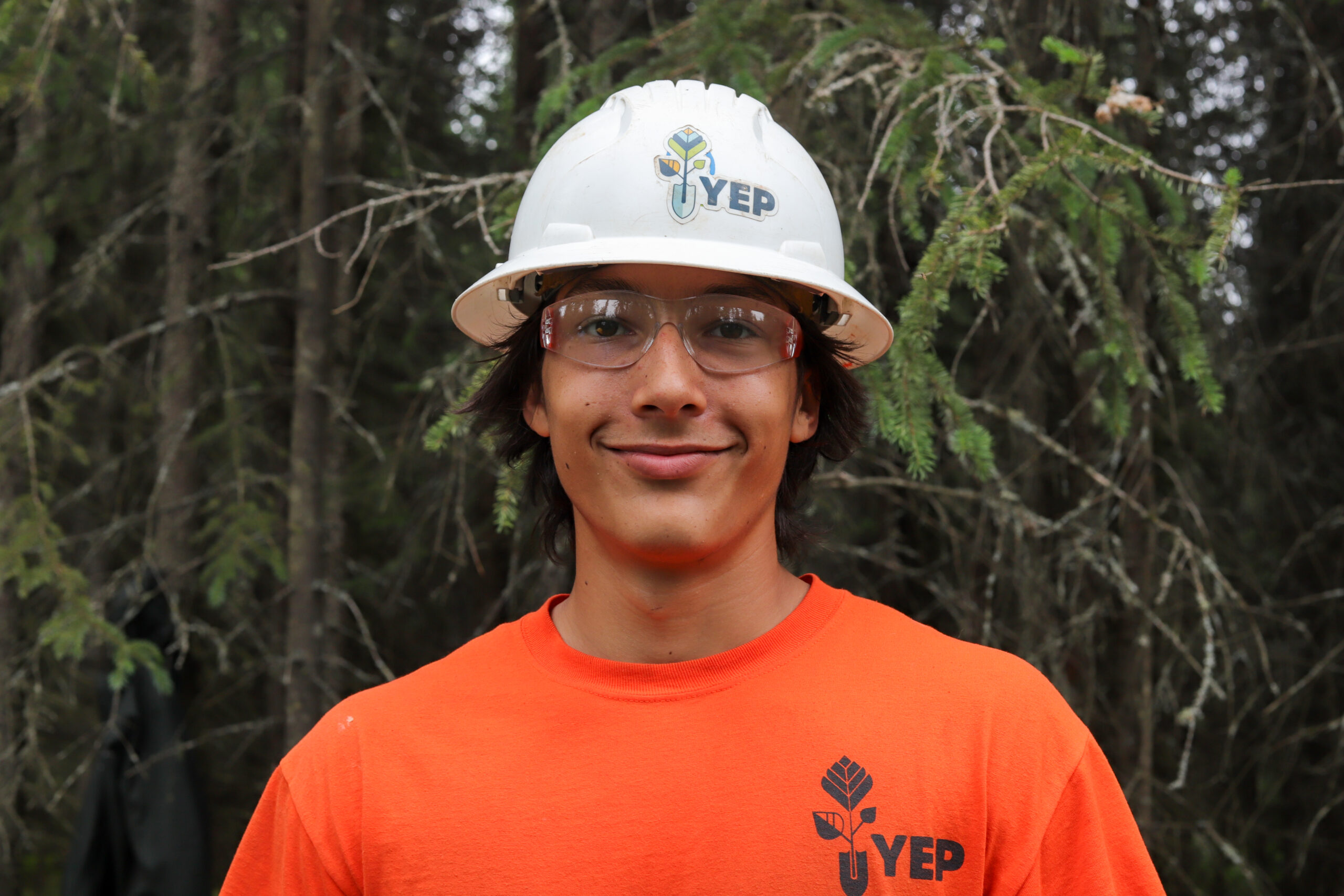 Close up of a person with an orange shirt, safety glasses, and a hardhat in the woods.