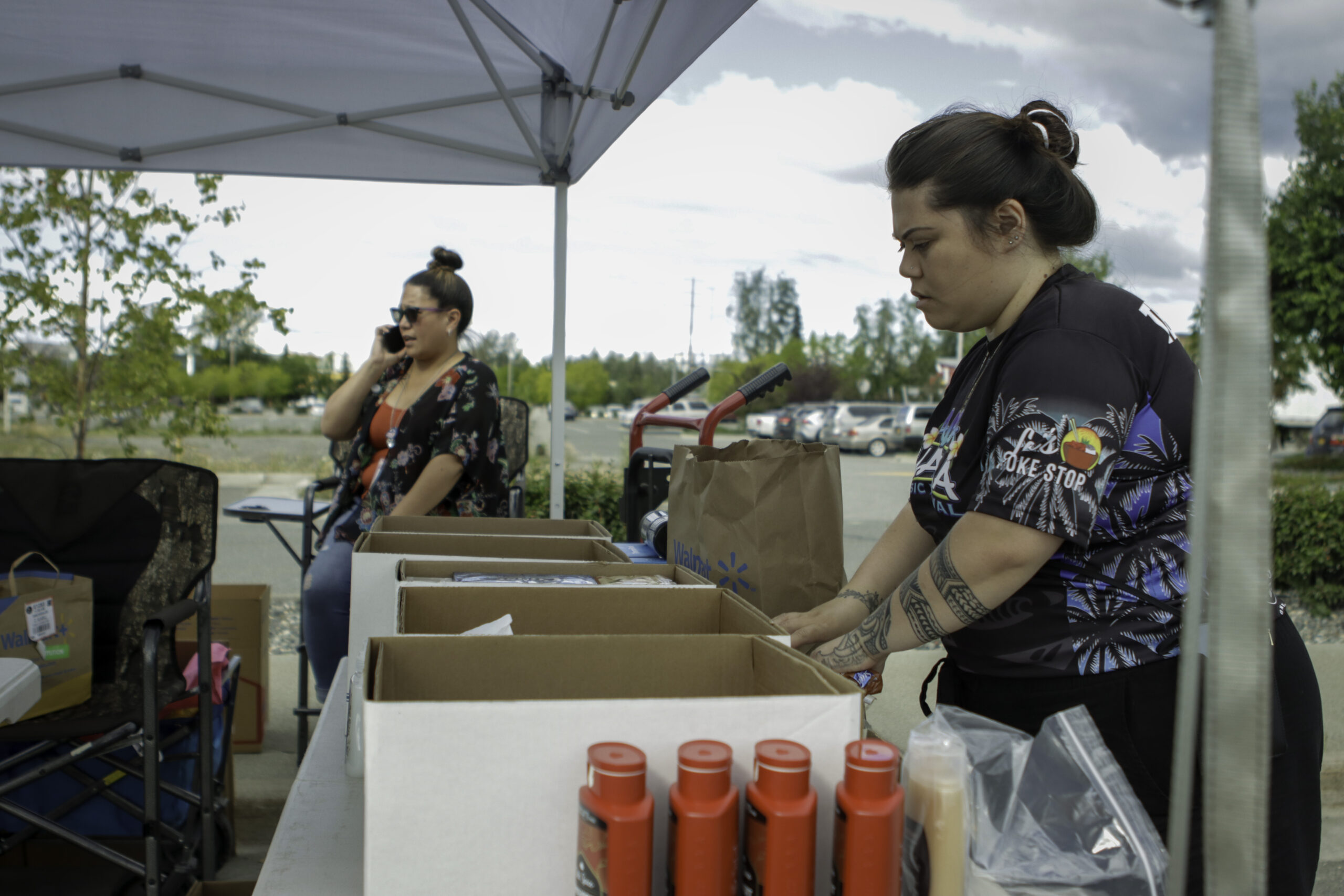 The woman on the right with a black shirt is putting shampoo bottles into a box. The woman on the left is sitting on a folding chair talking on a mobile phone.