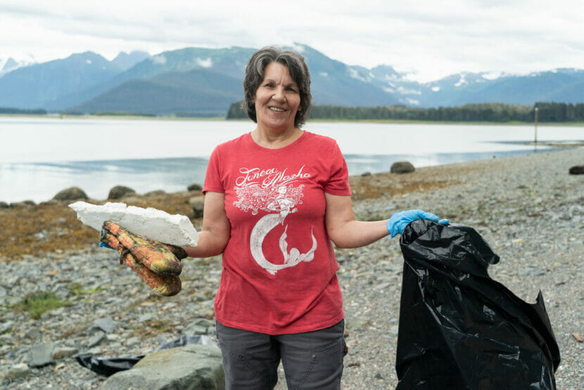 a woman stands on the beach holding a trash bag, oven mitts and a piece of styrofoam