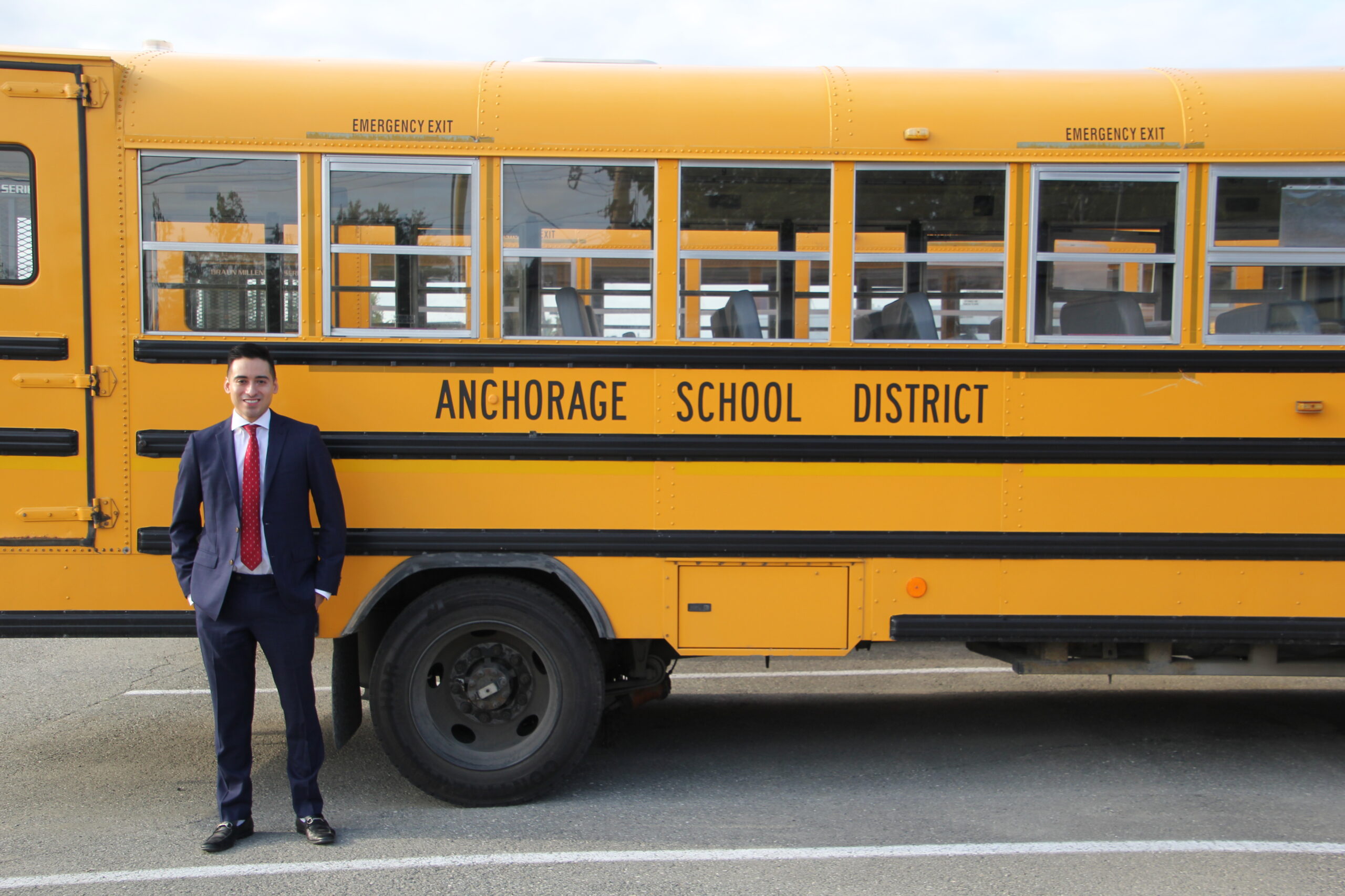 A man stands in front of a yellow Anchorage School District bus wearing a suit with a red tie.