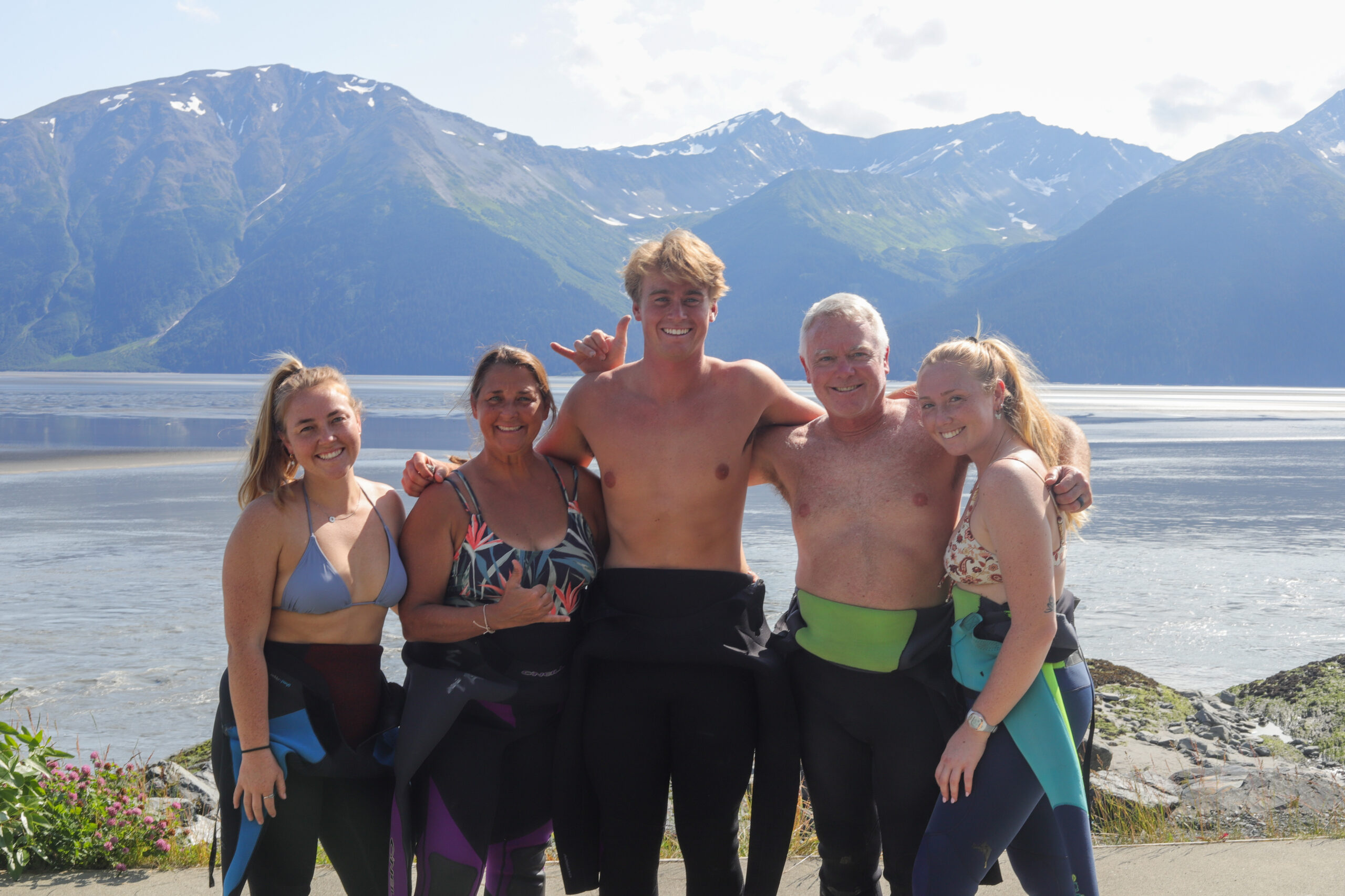 A group of people in wetsuits smile and do the "hang loose" gesture.