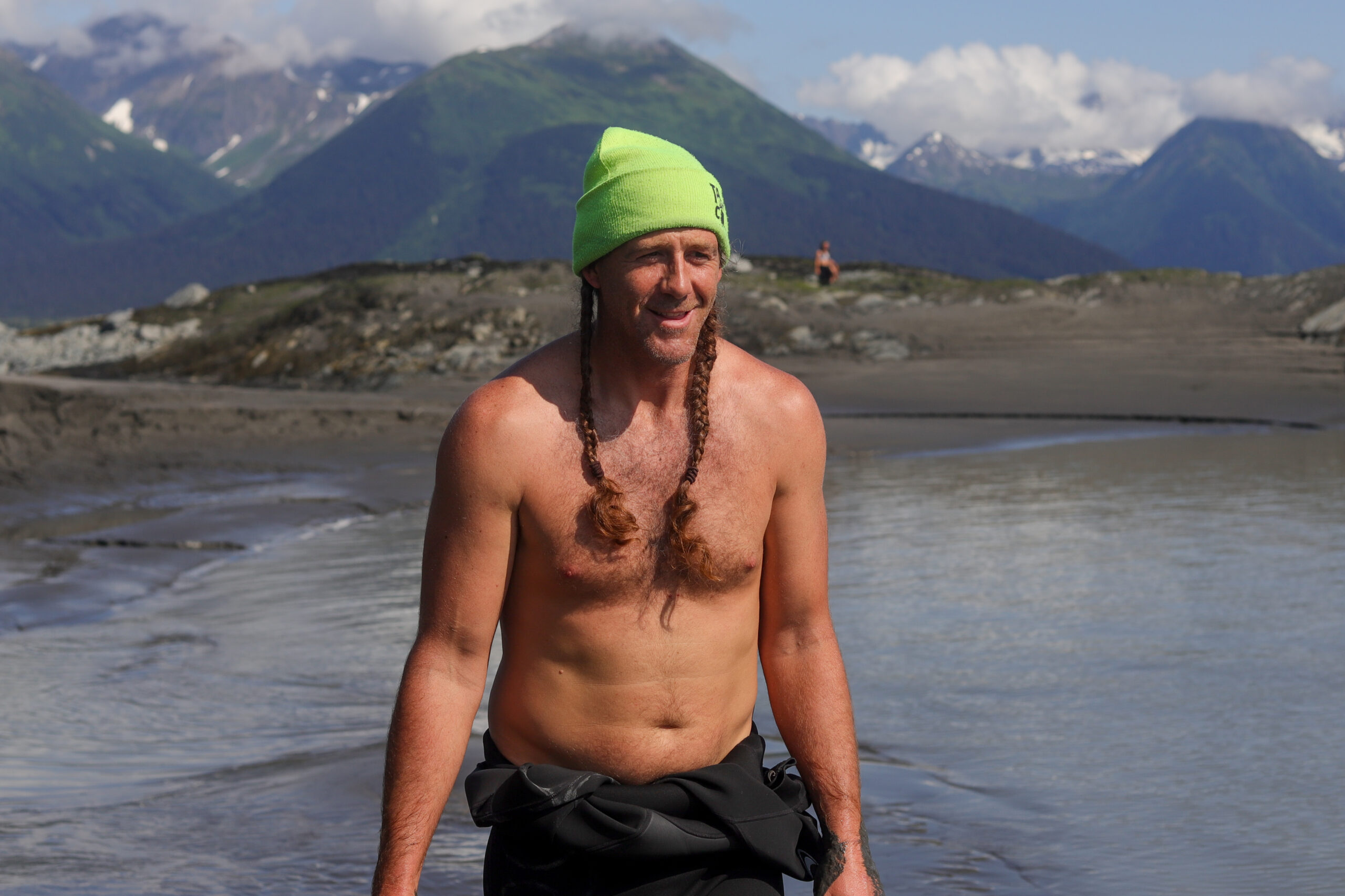 A man with a beanie, no shirt, and a wetsuit tied around his waist stands in a body of water.