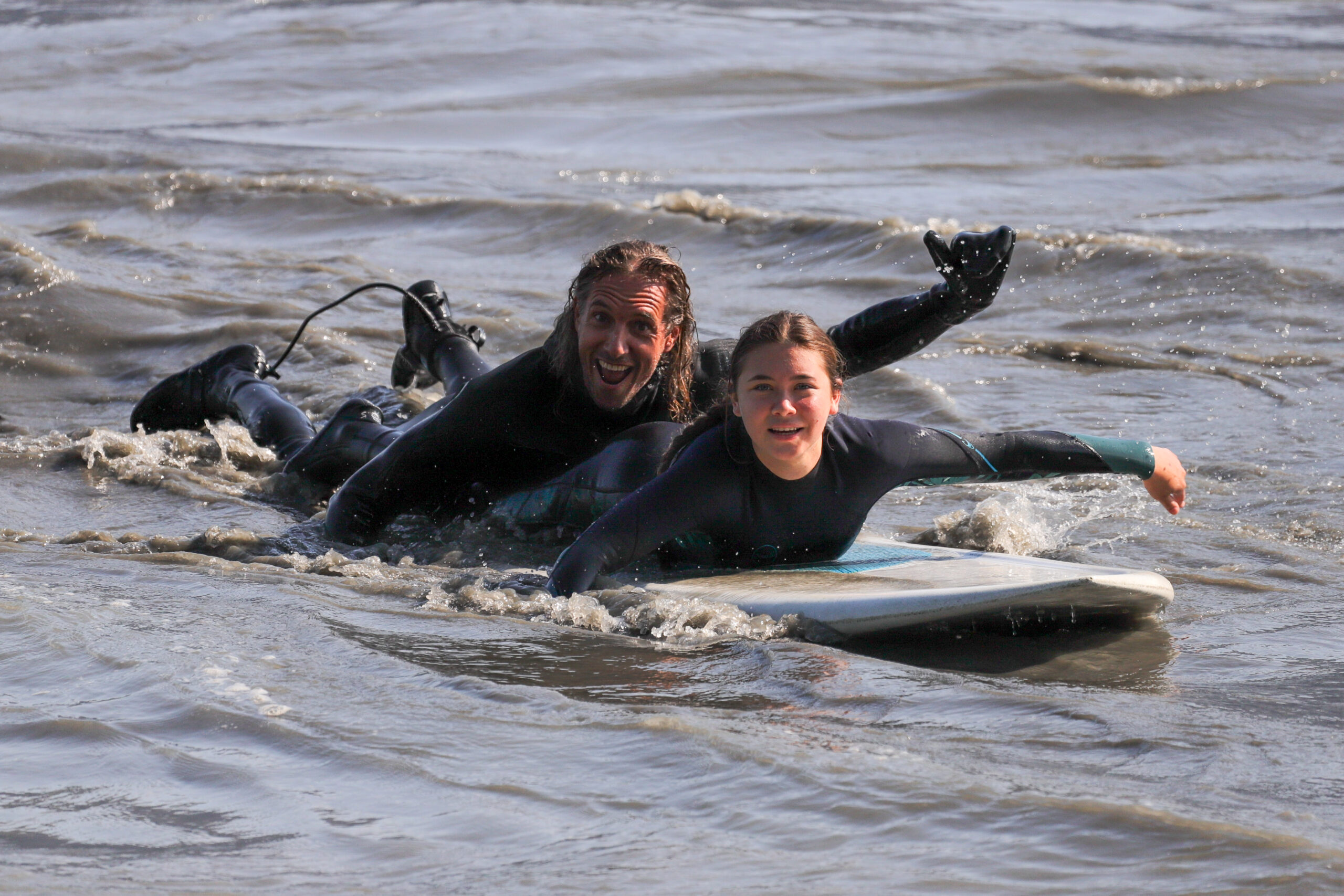 Two people ride on the same surfboard. One is giving the "hang loose" sign.
