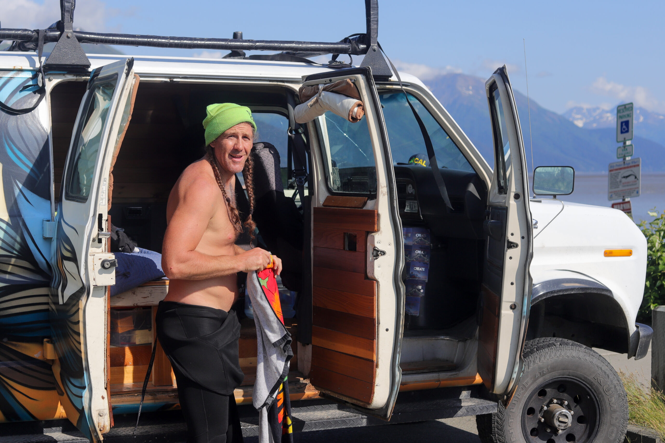 A man in a wetsuit and beanie stands next to a van, holding a towel.
