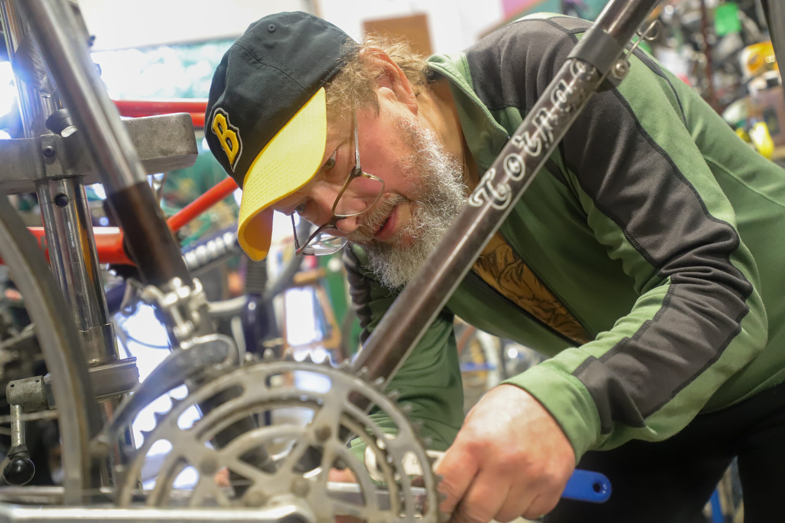 A man with glasses and a beard leans over a bike, and looks at its gears.