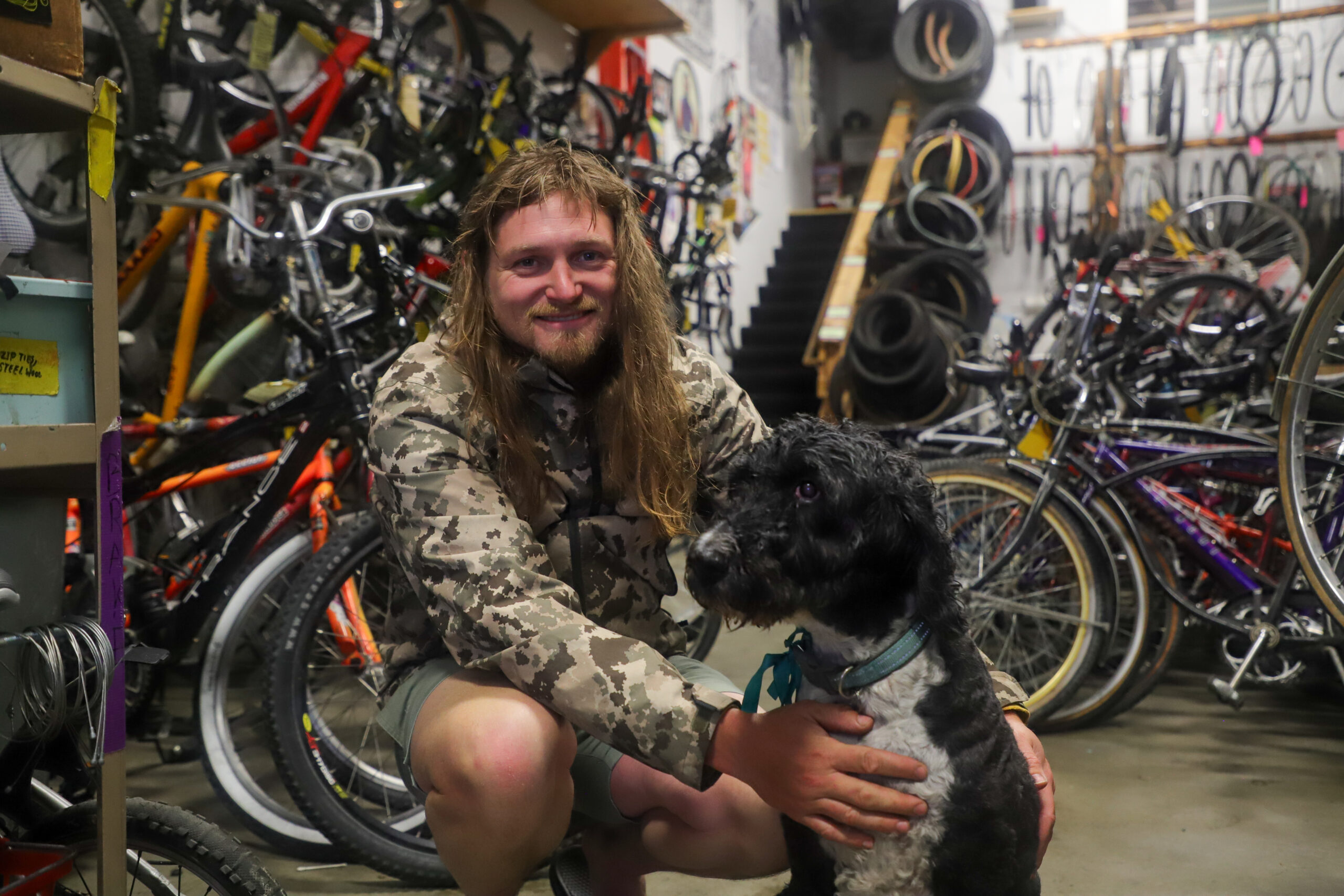 A man with long hair sits next to a black dog in a room filled with bikes.