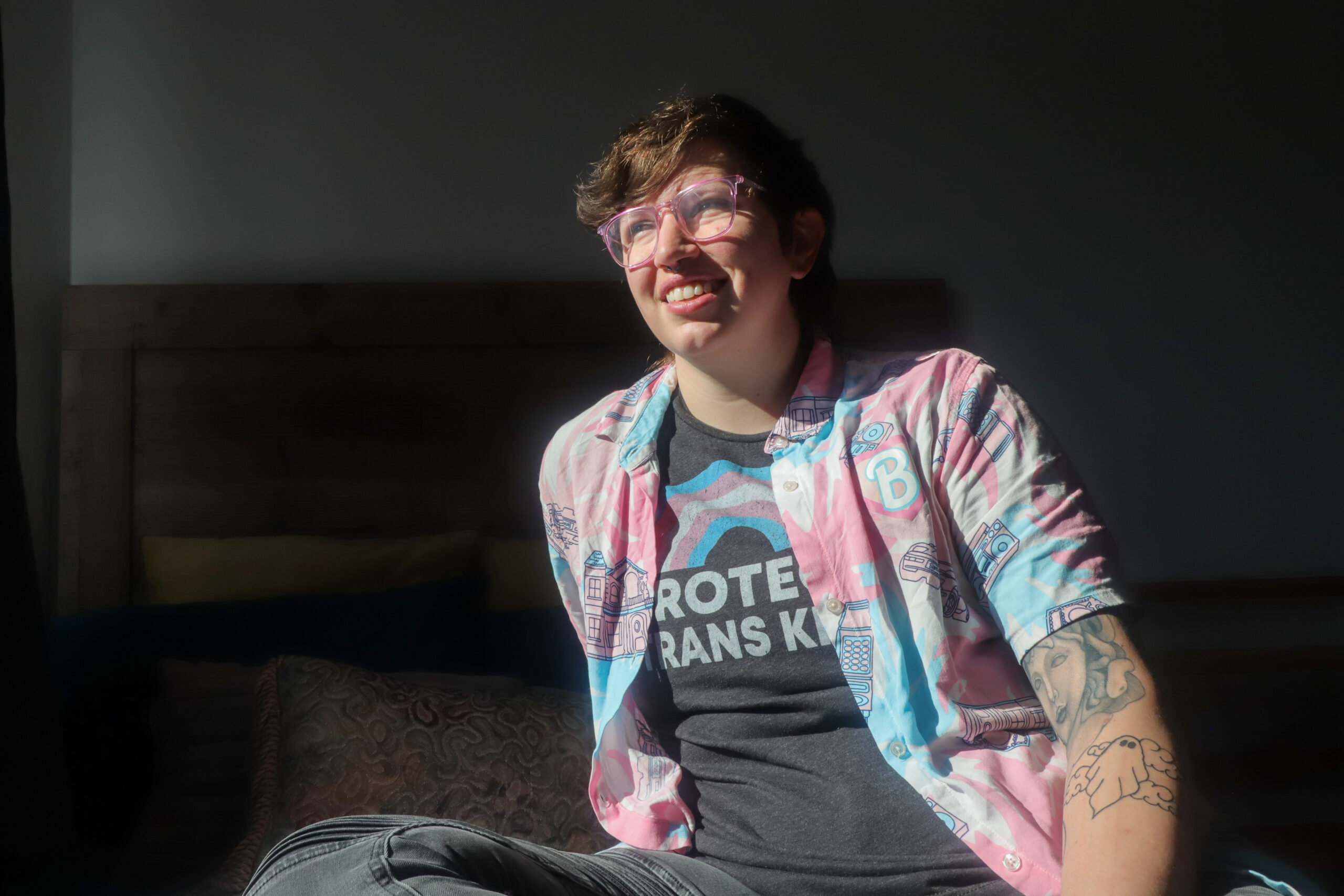 A person with glasses, a pink and blue collared shirt, and a t-shirt that reads "PROTECT TRANS KIDS" sits on the edge of a bed.
