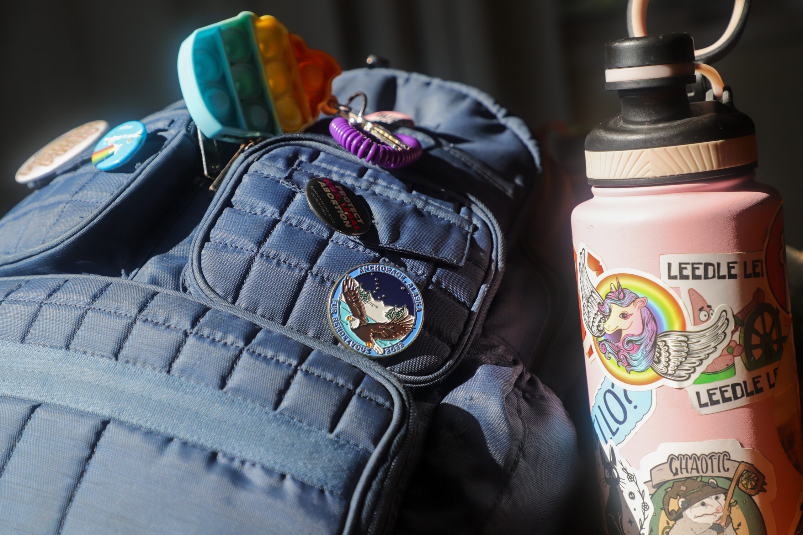 A blue backpack covered in pins next to a pink water bottle covered in stickers