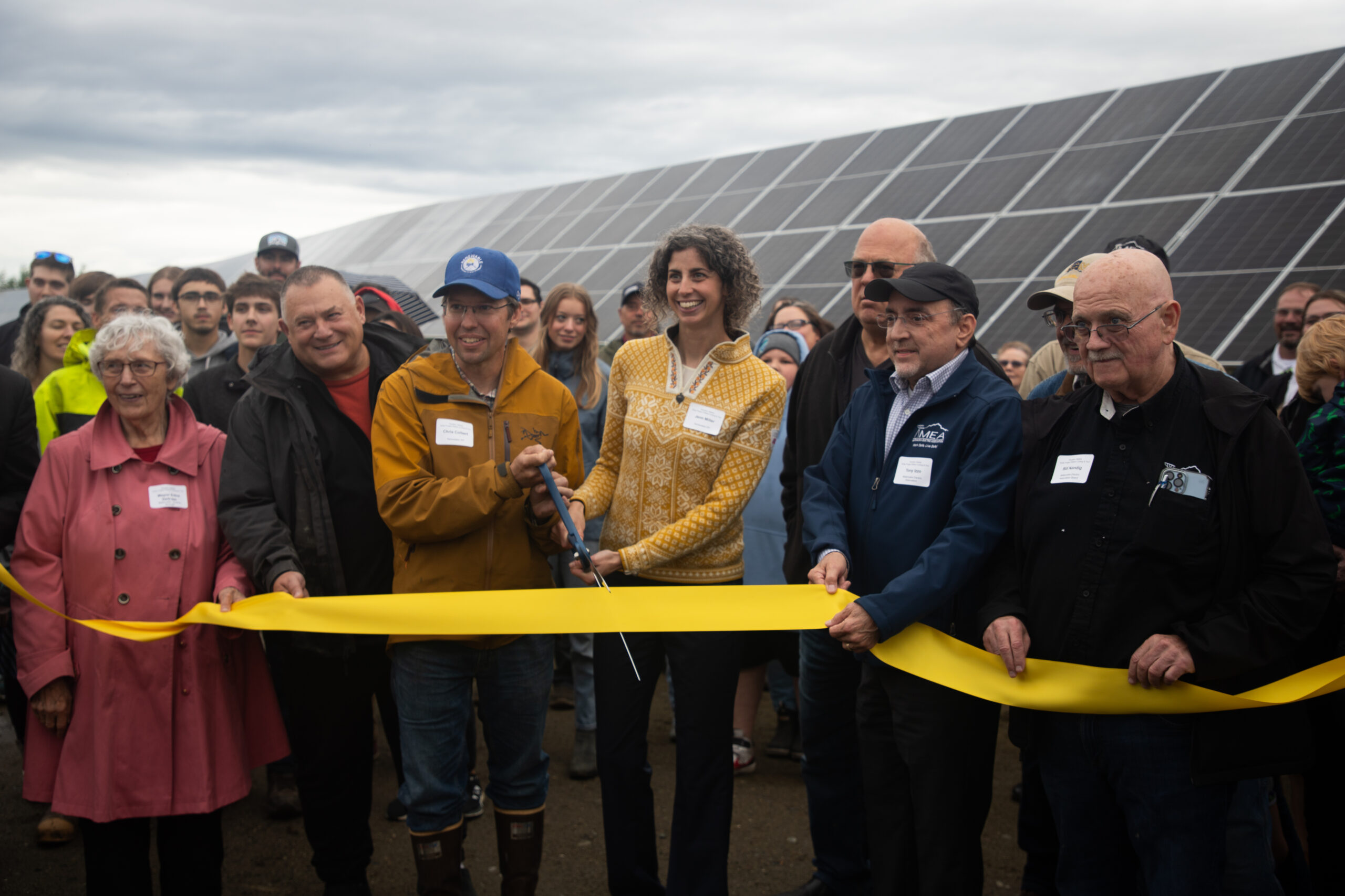 A crowd of people gather in front of solar panels as those in the front hold a yellow ribbon that is about to be cut by two people holding large scissors.