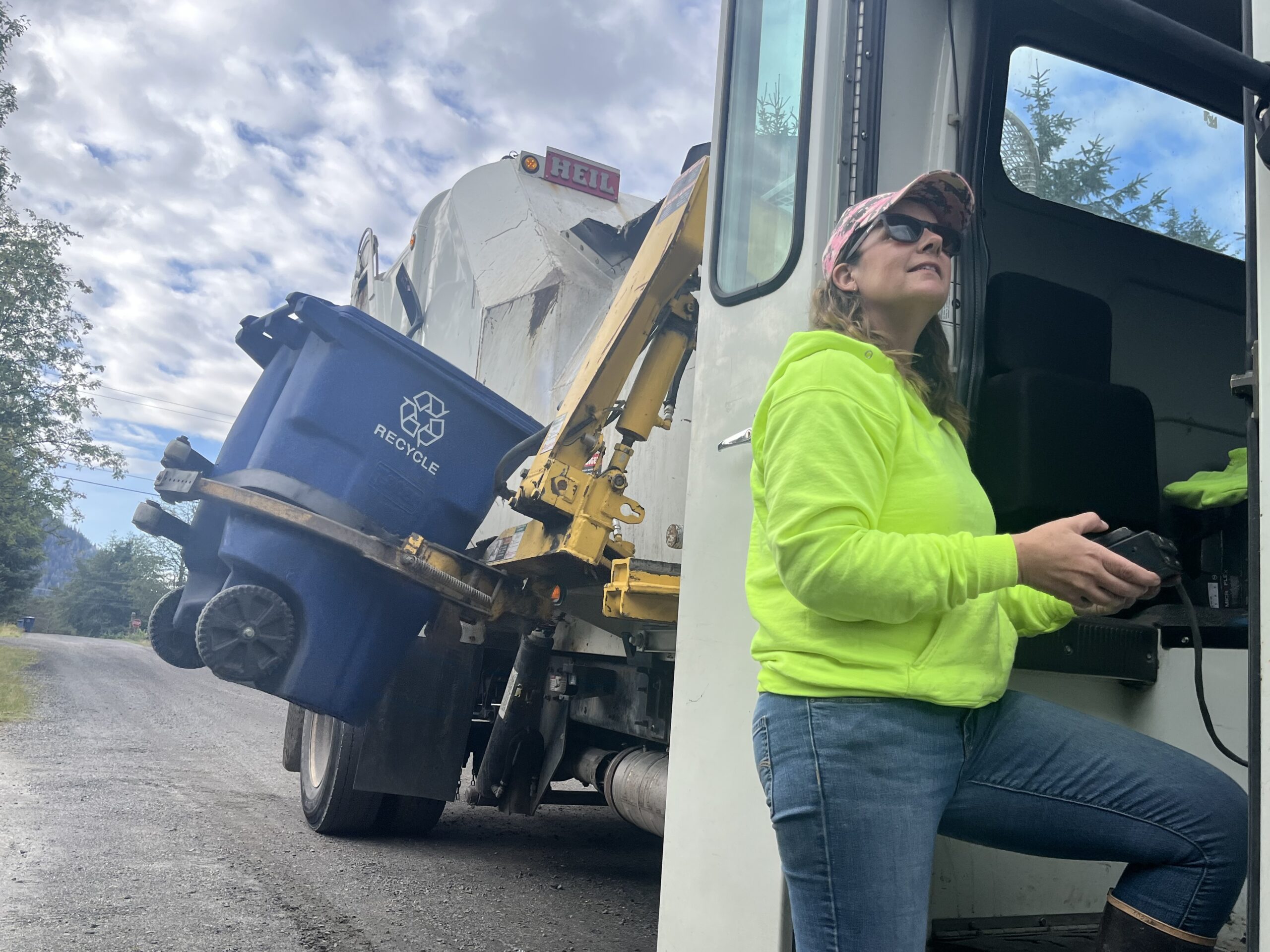 A woman in a bright neon hoodie operates a trash truck's mechanical arm, to pick up a recycling bin