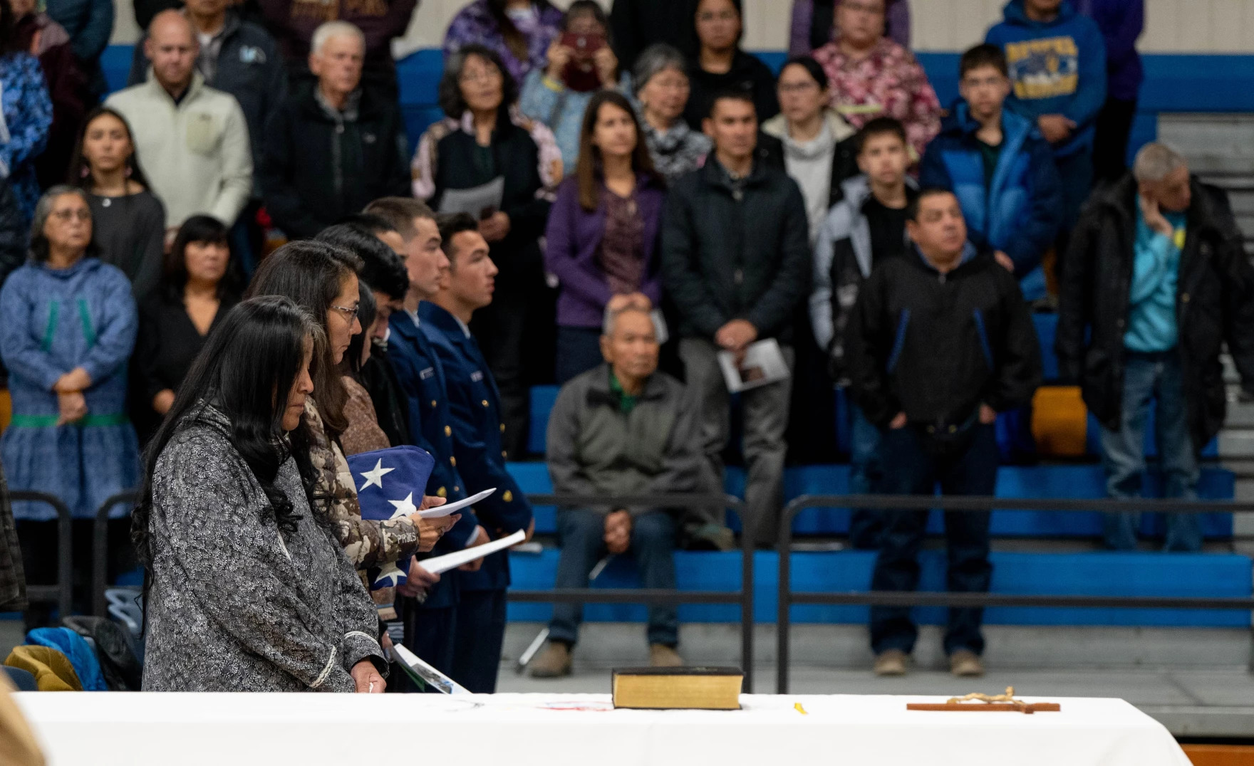people stand in a line, near a casket, as a crowd looks on in a school gym
