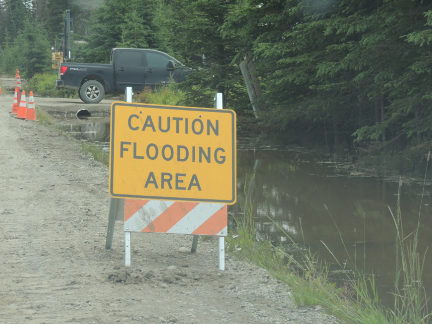A yellow sign says "caution flooding area" near a flooded ditch.