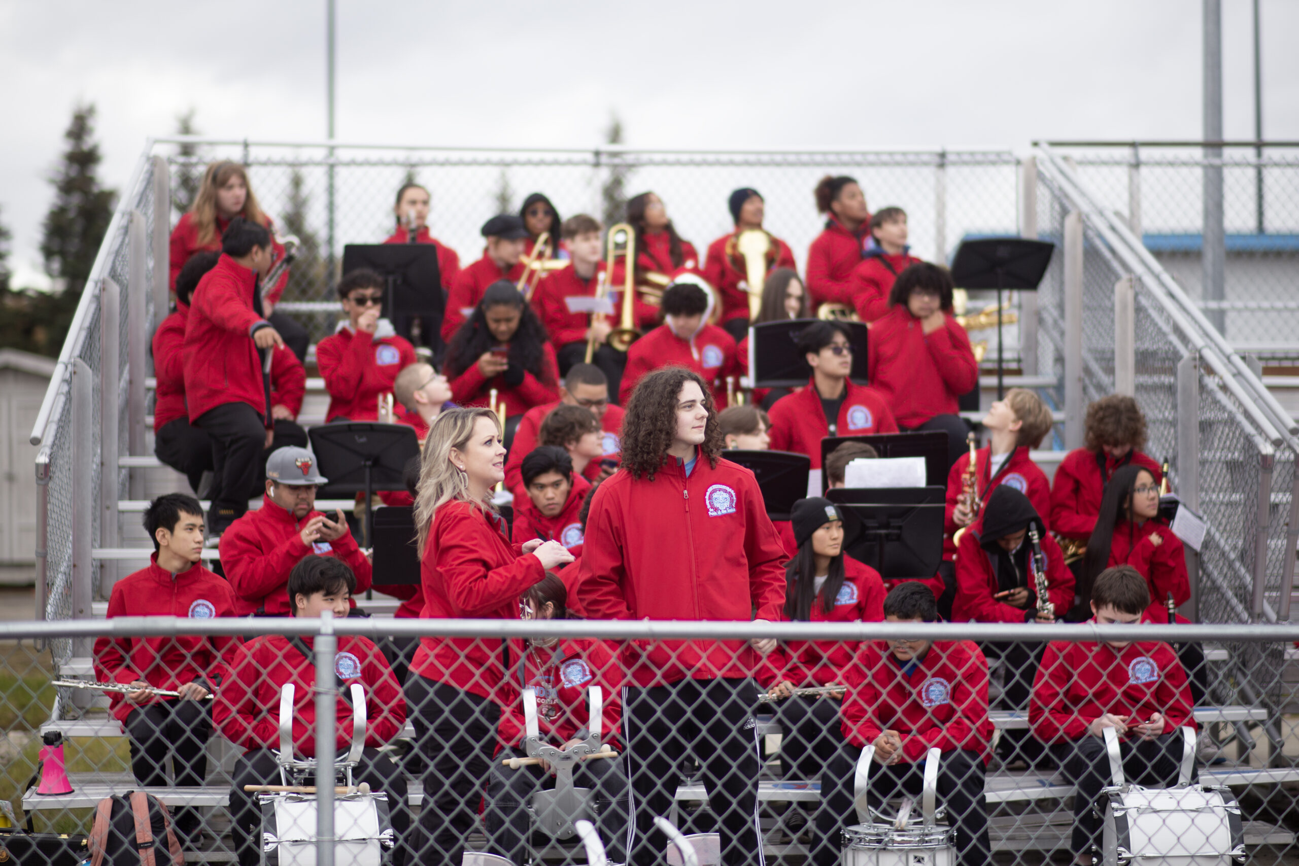 A marching band in matching red jackets sits in the bleachers of a football game between Bettye Davis East High and Bartlett on Sept. 30, 2023 (Shiri Segal/Alaska Public Media)