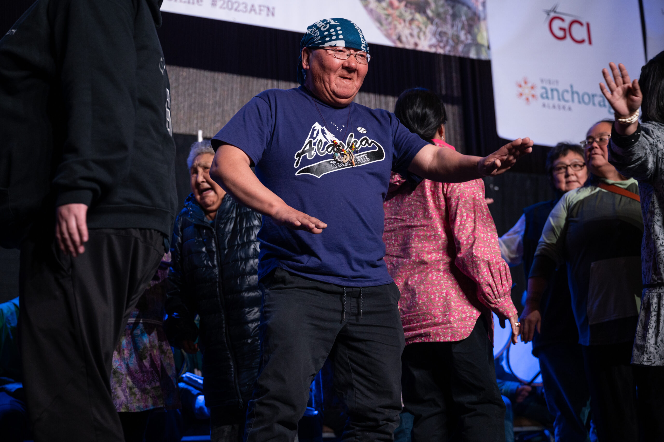 a man with a bandana on his head dances on stage with a group of Alaska Native Dancers.