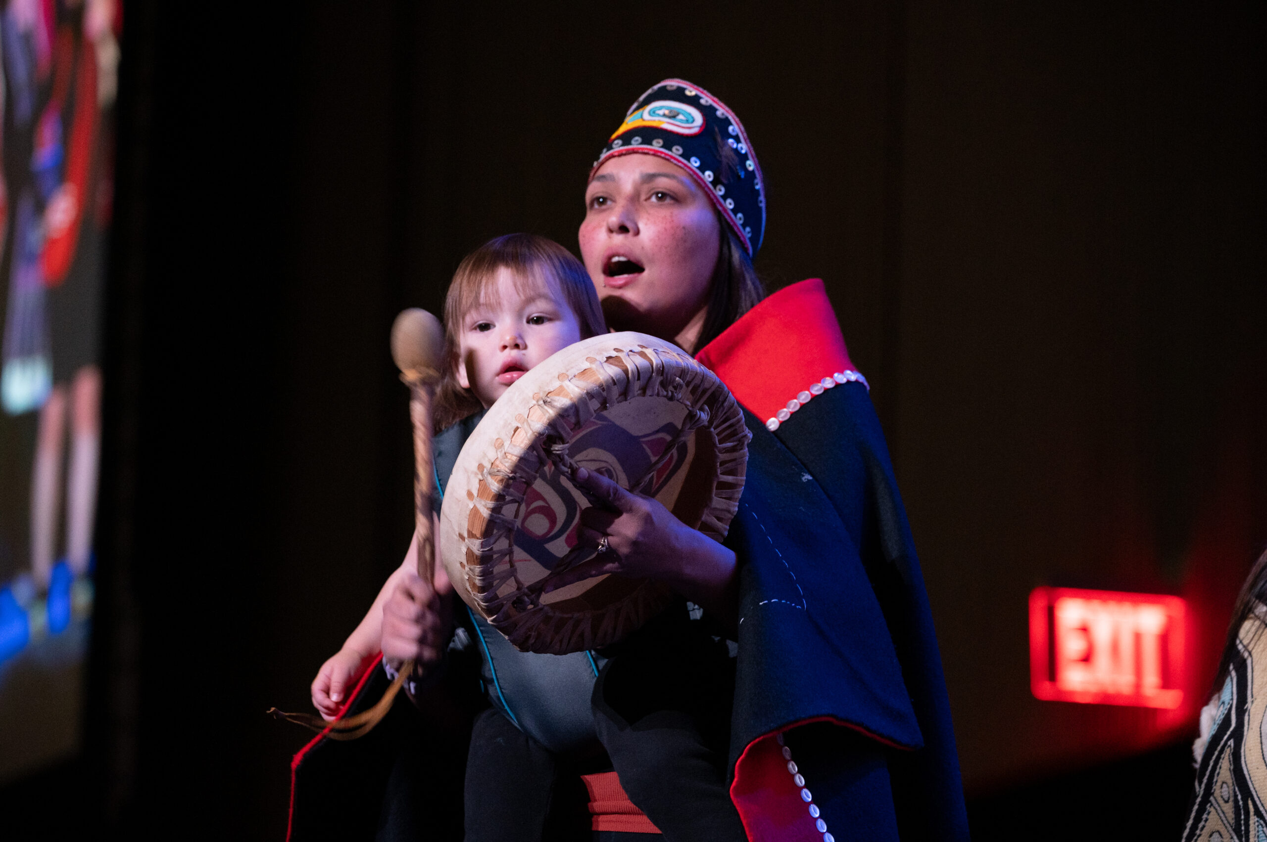 A woman in Alaska Native clothing holds a drum while dancing with her newborn.