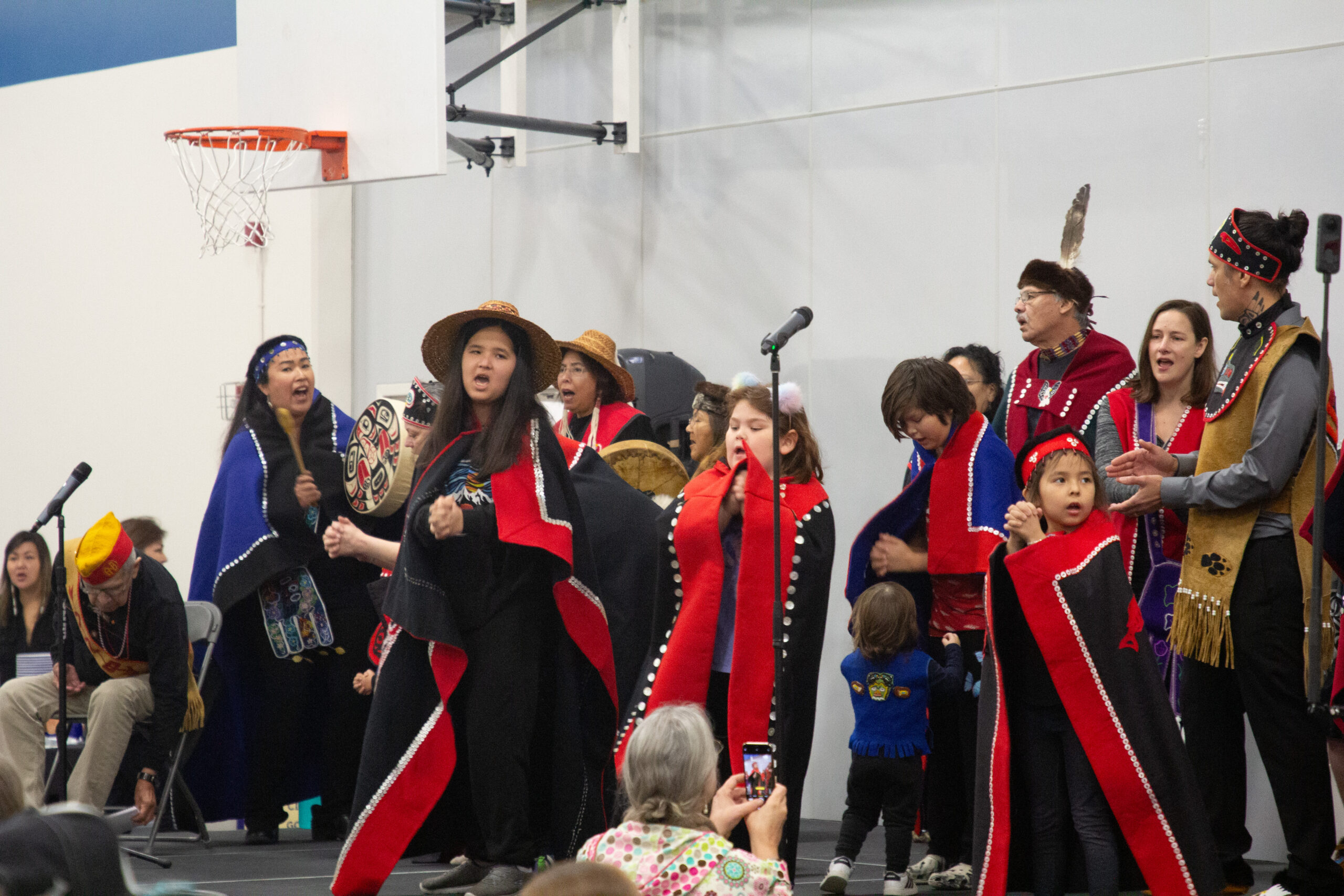 A group of dancers sing before a crowd on stage. Some holding drums, others clasing their hands together while wearing traditional Tlingit attire.