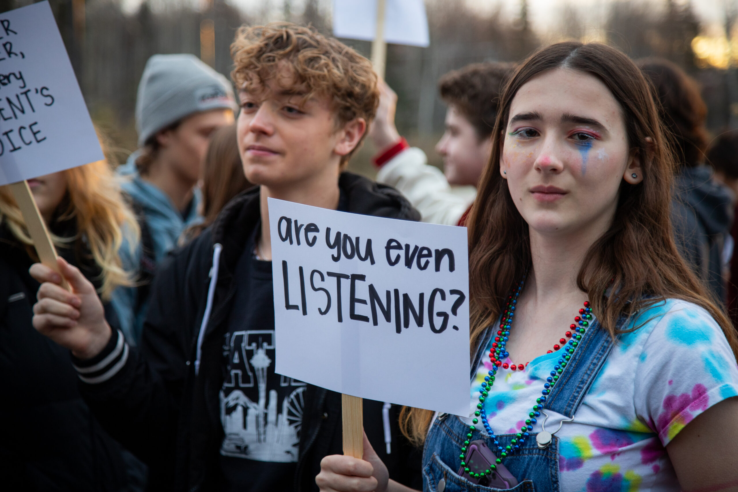 A student wearing colorful makeup holds a sign that says "Are you even listening?" during a walkout protest at Career Tech High School.