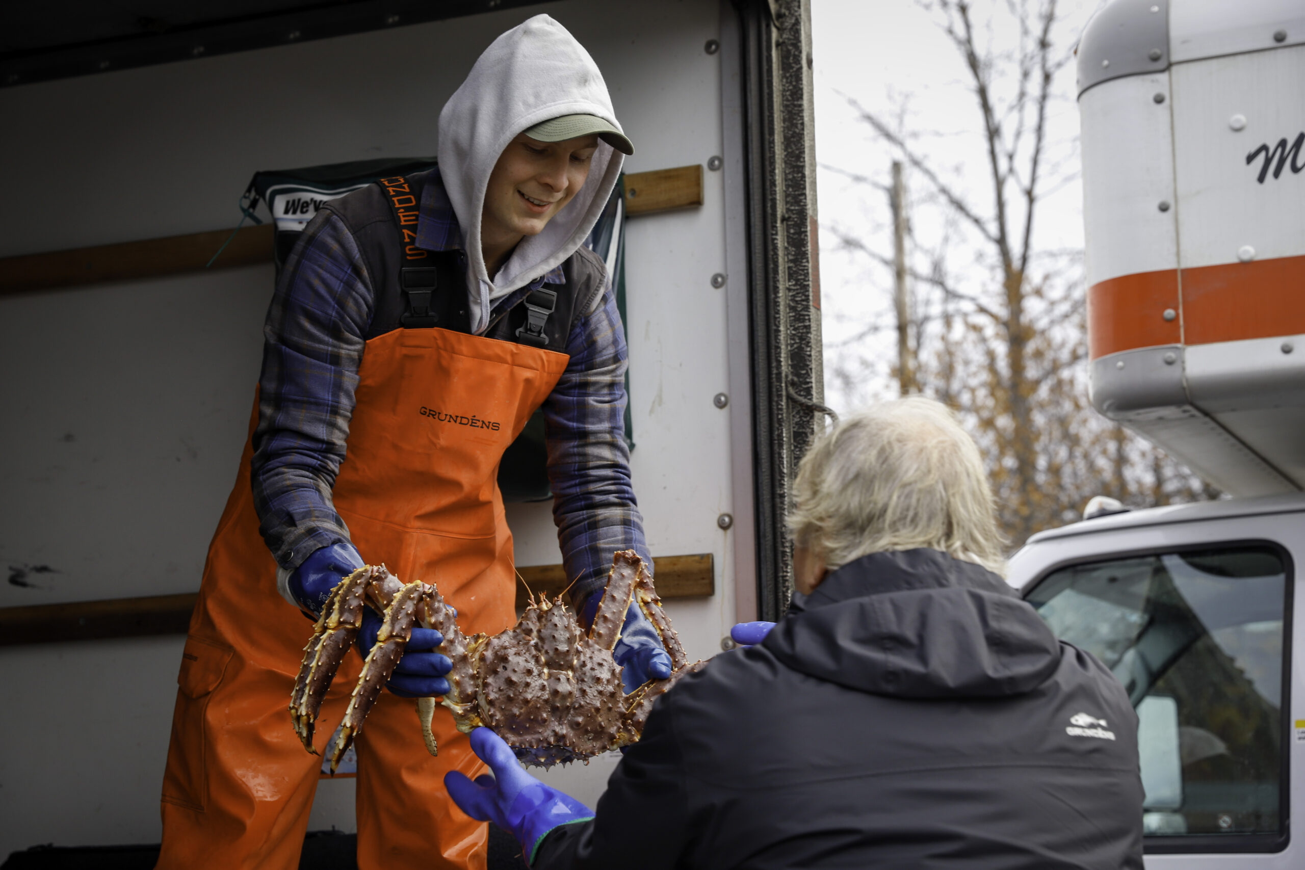 A man with orange waders and a hoodie hands over a live crab to an old man