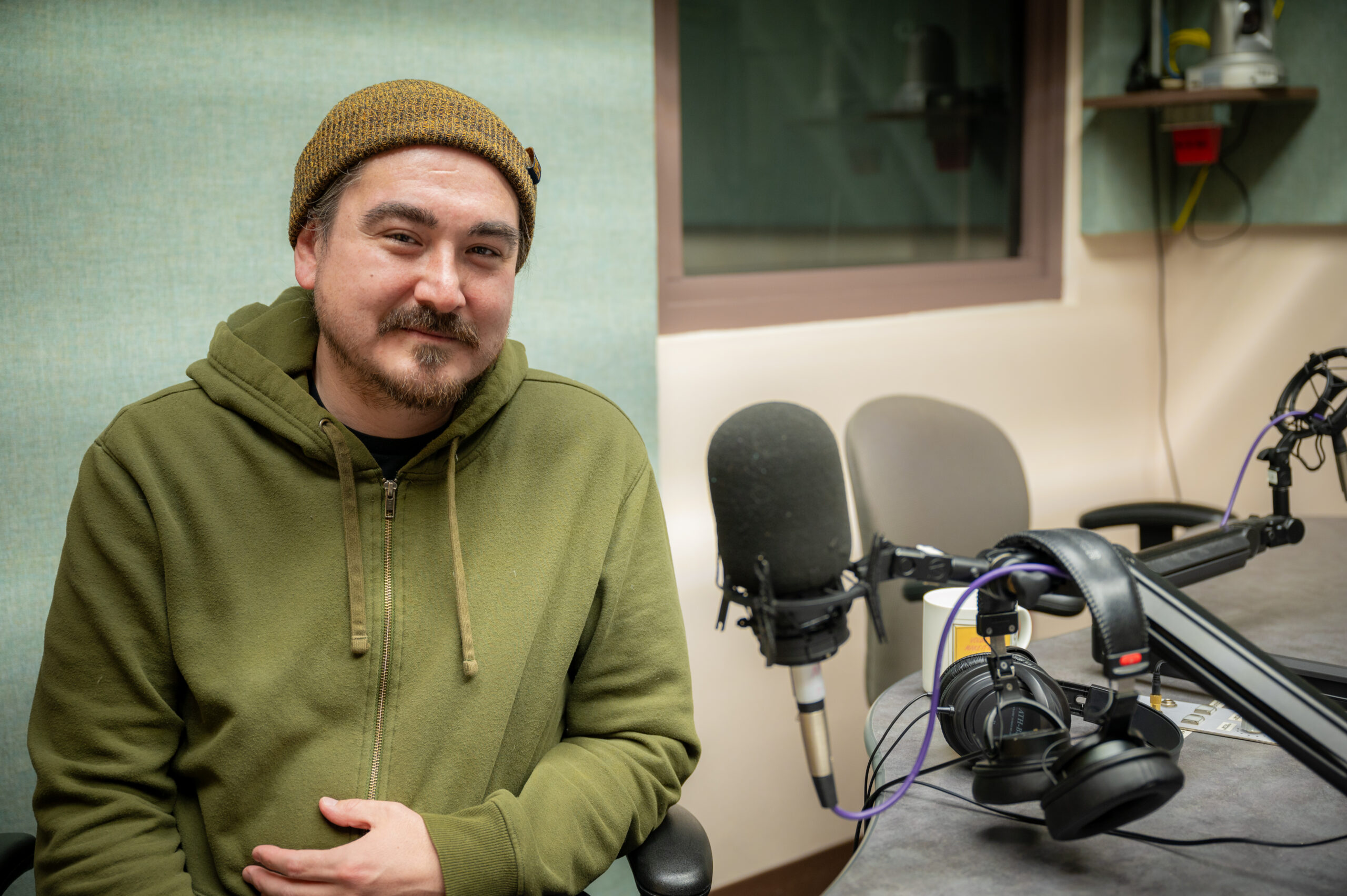 a man with a beanie hat on sits by a studio microphone.