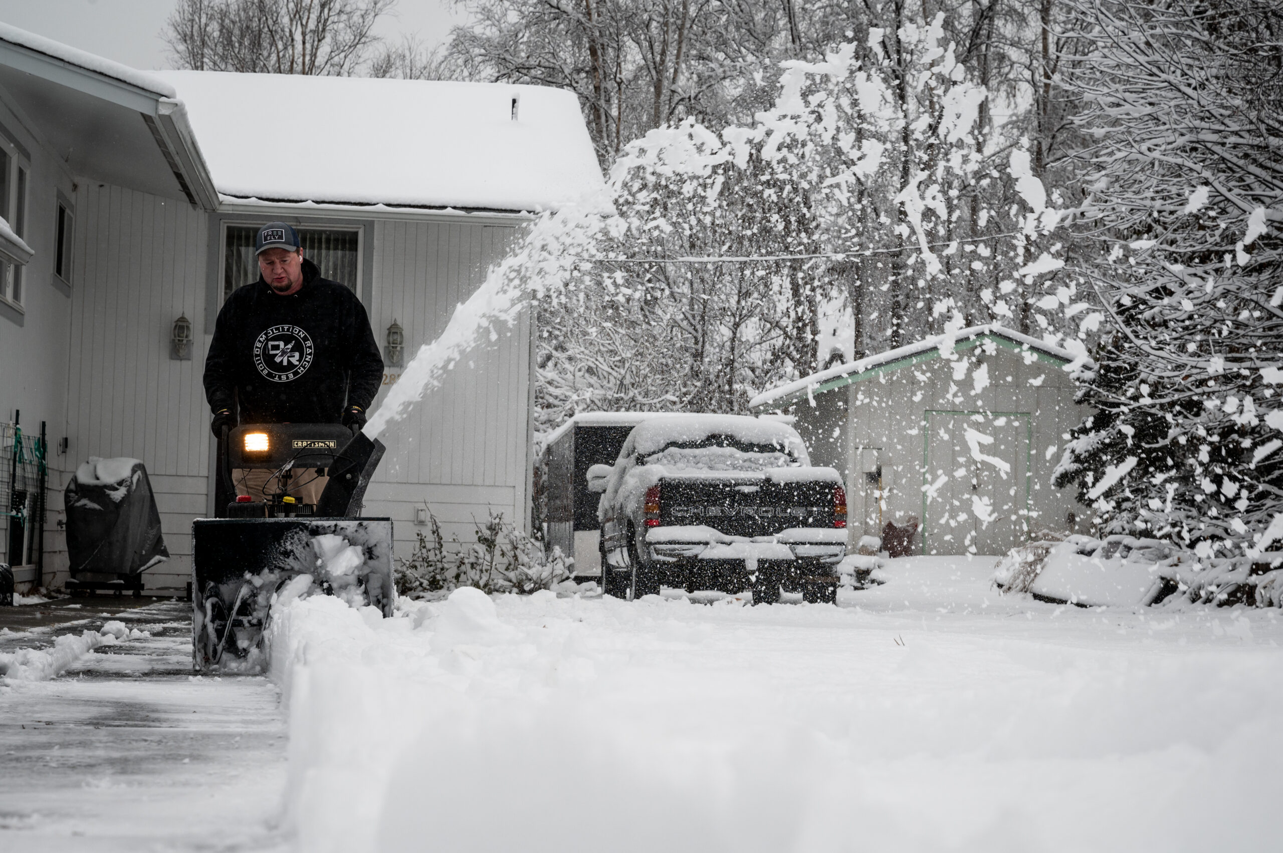 A man in a black hoodie uses a snow blower on his driveway.