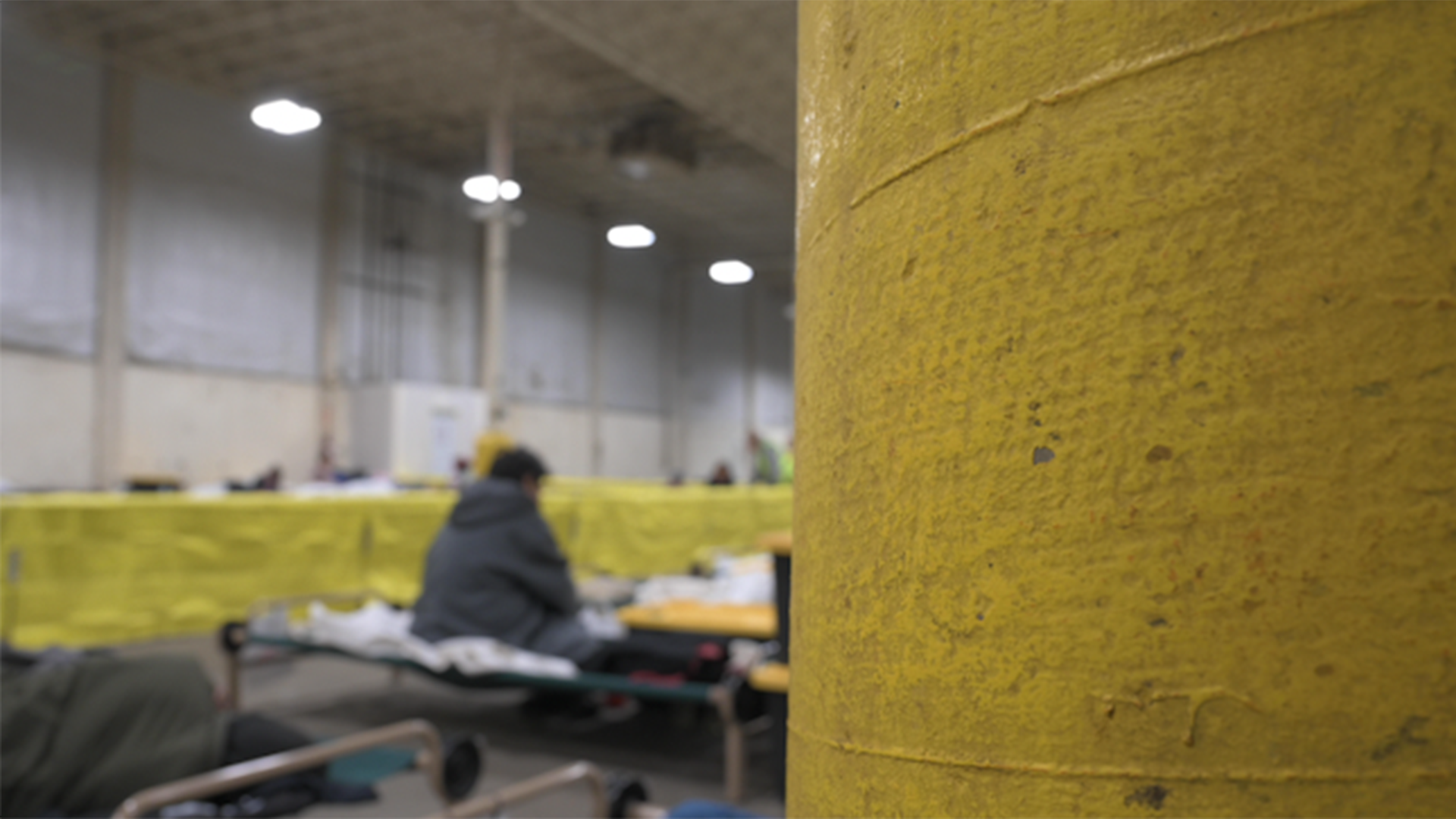 A man in a dark green jacket sits on a cot alone inside a building that has been converted into a shelter.