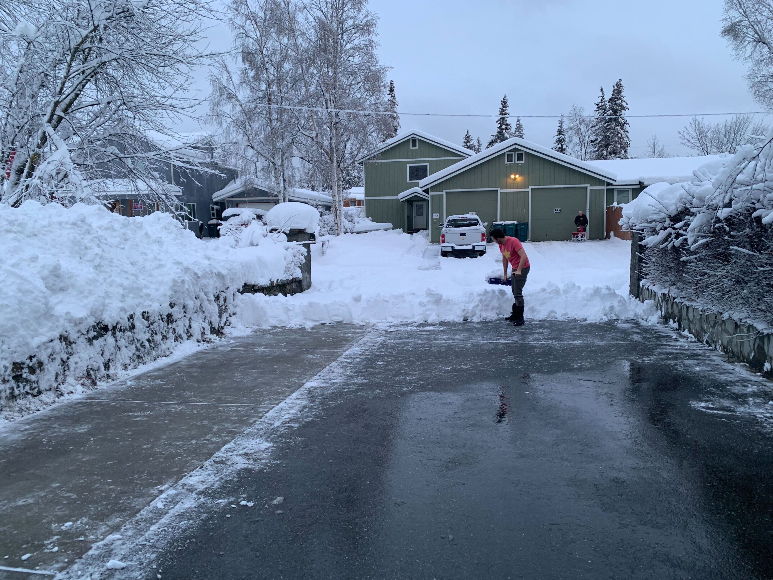 A man shovels snow from his driveway late in the day in front of a green house.
