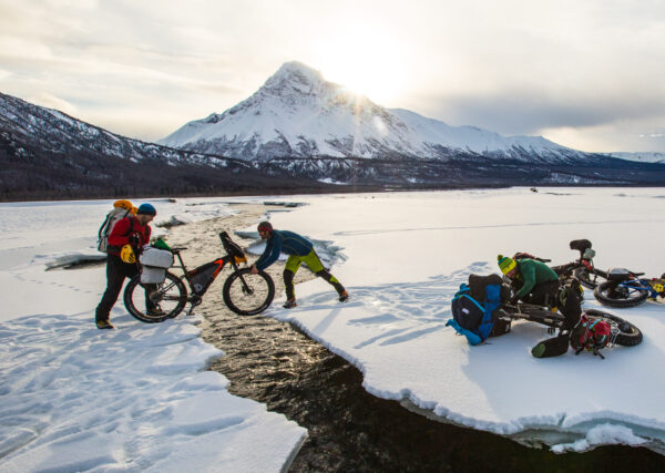 People pass a fat tire bike across open water while standing on ice. Other bikes sit in the snow.