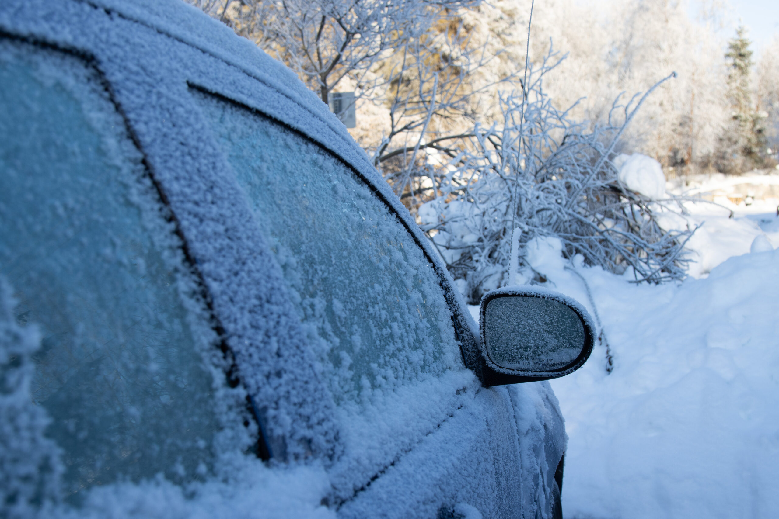 A car covered in frost sits in the shade with snow covering the ground.