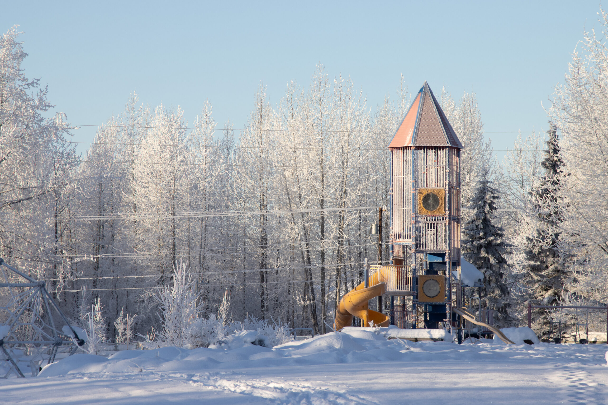 A children's playground covered in frost and surrounded by snow with sunlight shining on it.